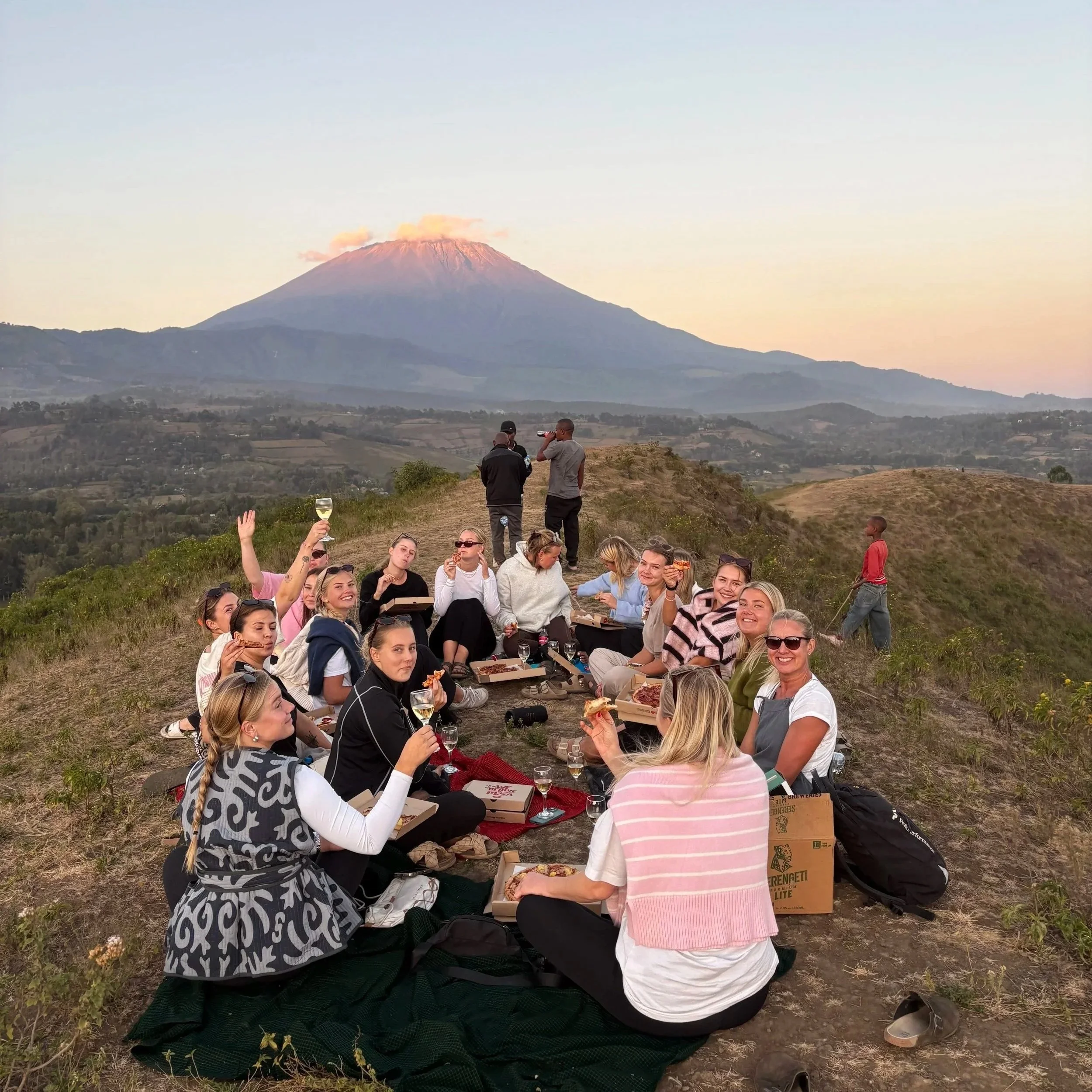 Grupp människor, som sitter på marken och äter pizza, med vulkanen Mount Taranaki i bakgrunden