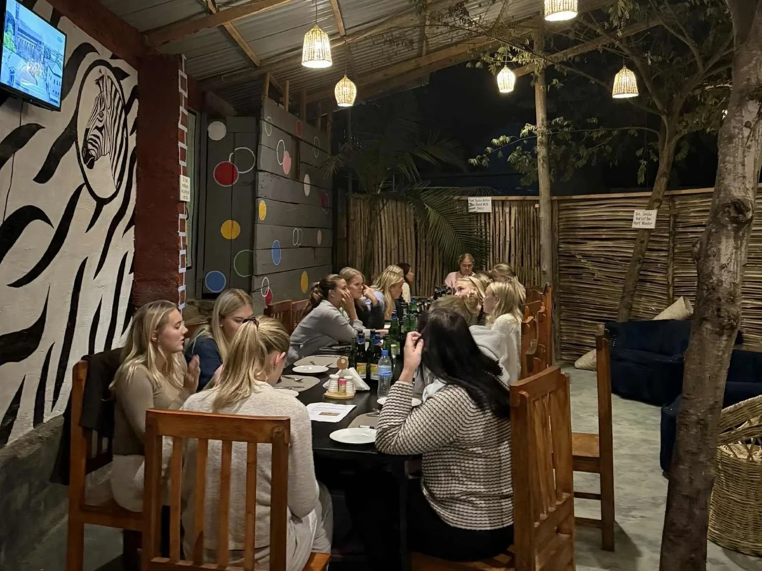 Många personer som sitter vid ett bord i en restaurang eller kafé utomhus, omgiven av väggar av trä och dekorativa mönster, under varmbelysta hängande lampor.