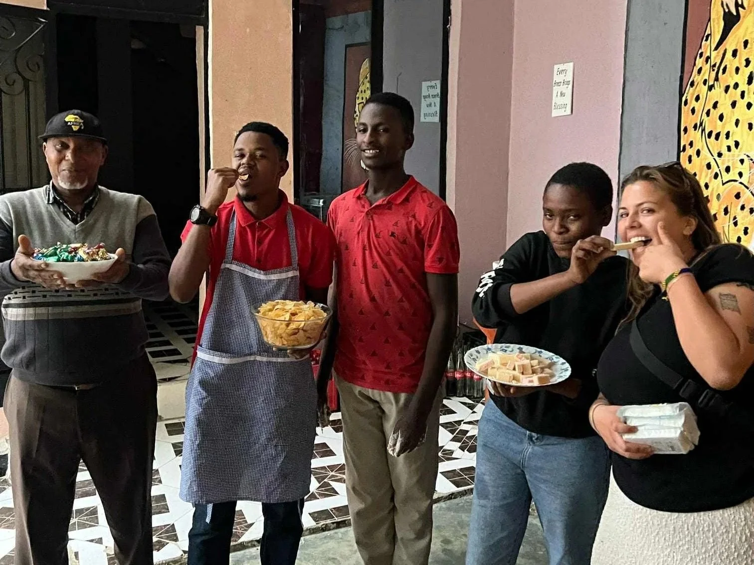 Fem personer som står i en rad och äter snacks, inklusive chips och godis, i ett inomhusmiljö med konstnärlig väggdekoration.