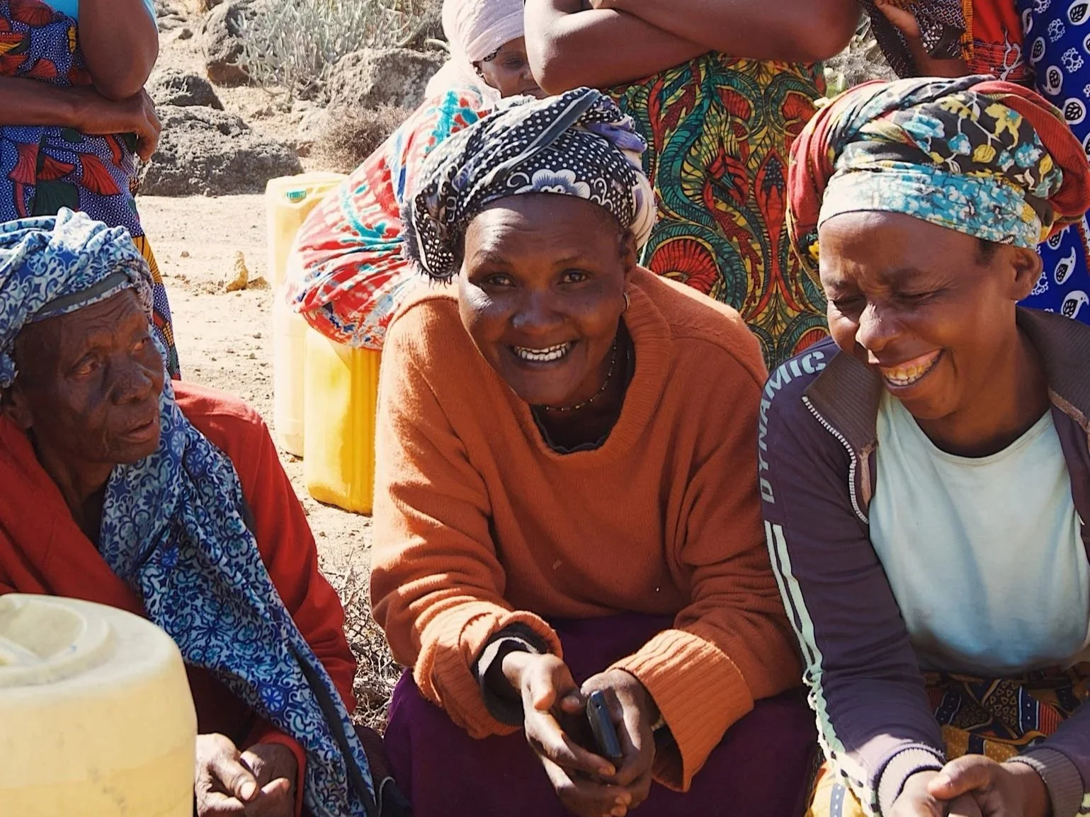 En grupp kvinnor med färgglada kläder och huvuddukar, som verkar skratta och umgås utomhus på en solig dag.