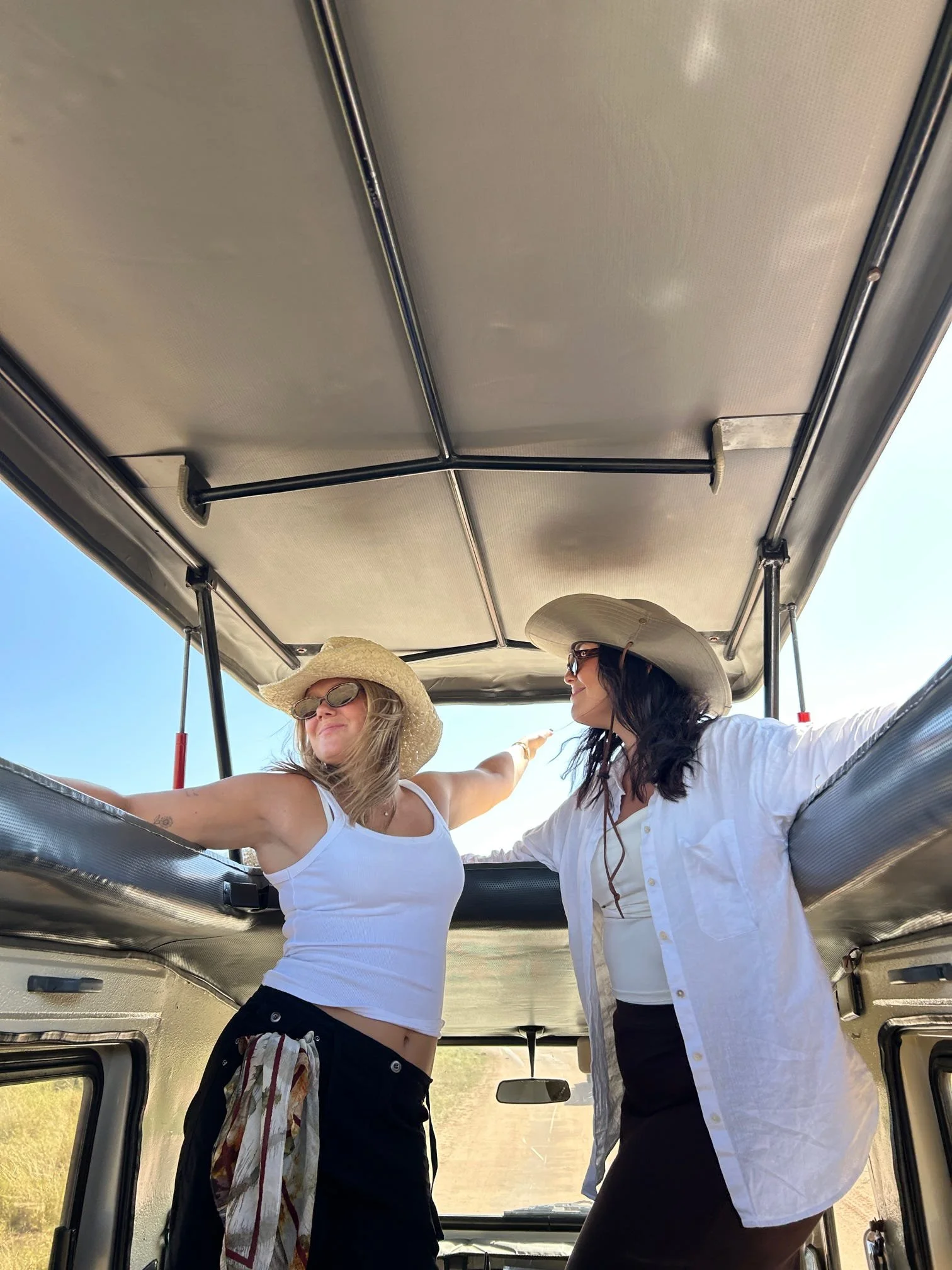 Två kvinnor i solhattar och solglasögon står i en öppen ledbuss och njuter av solen under en klar blå himmel.