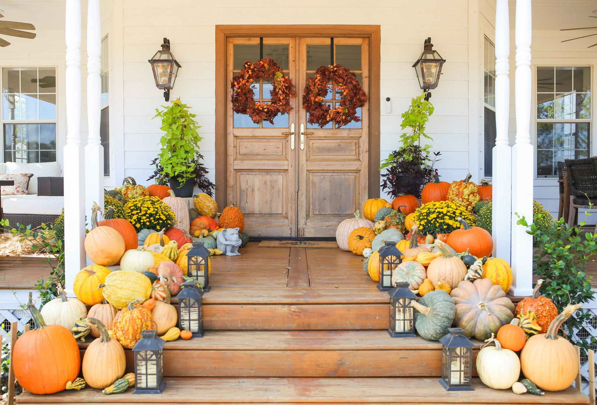 Fall porch decorated with pumpkins, gourds, and lanterns in front of wooden double doors with autumn wreaths.