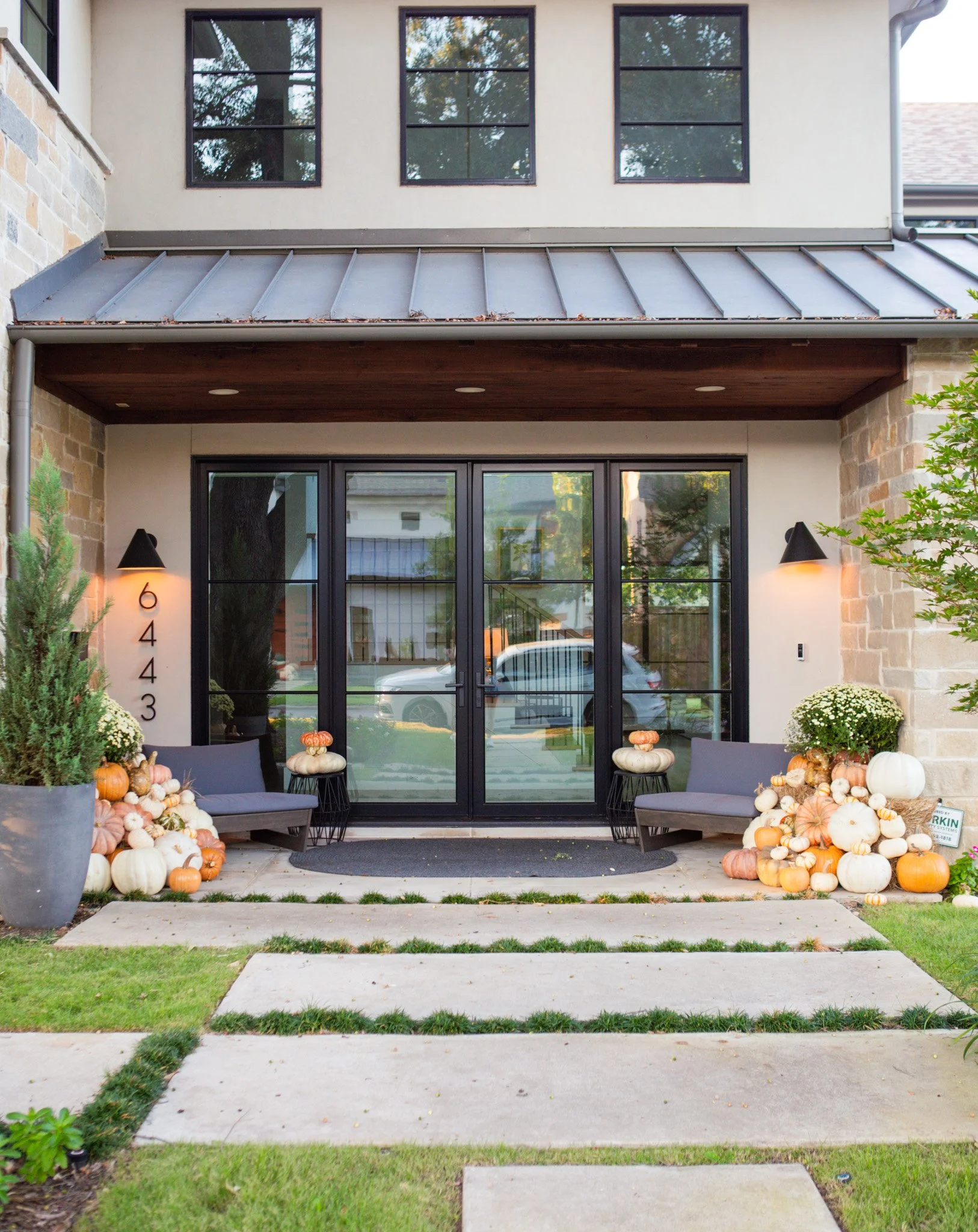 Front porch decorated for autumn with pumpkins, gourds, potted plants, benches, and wall-mounted lamps.