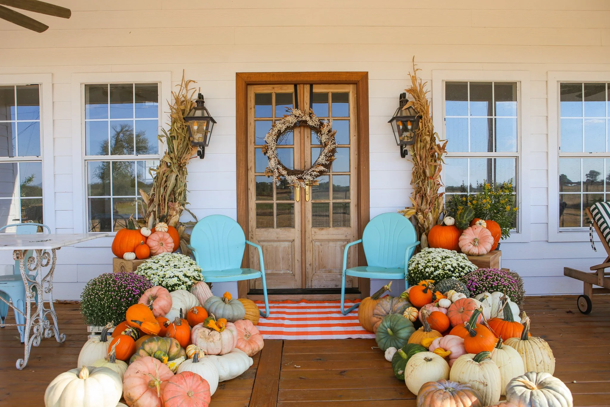 Front porch decorated for fall with pumpkins, mums, and blue chairs, featuring a wooden door with a wreath, lanterns, and dried corn stalks.