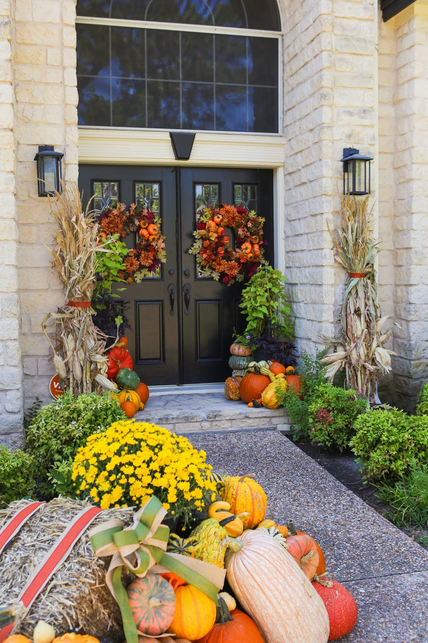Front entrance of a house decorated for fall with pumpkins, gourds, and autumn-themed wreaths on the black door. Cornstalks and greenery line the steps, with a yellow chrysanthemum in the foreground.