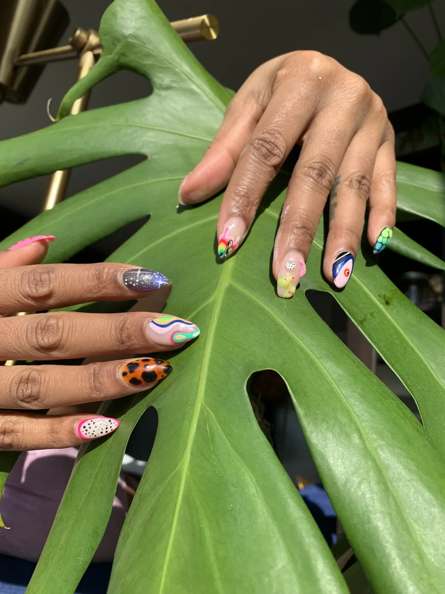 Close-up of people showing hands with colorful, patterned nail art, touching or resting on a large green leaf.