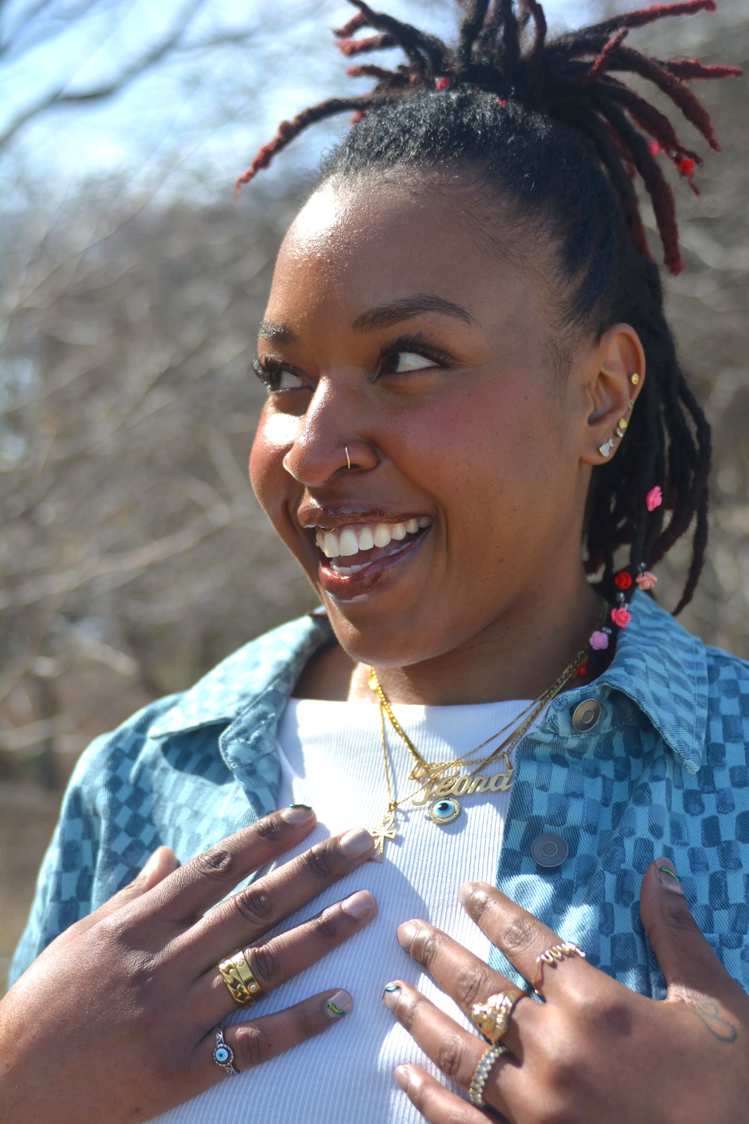 Close-up of a smiling Black woman with dreadlocks styled in a high ponytail and adorned with pink beads. She has multiple earrings, a nose ring, and wears gold rings, necklaces, and a blue patterned jacket over a white shirt. She places her hands on her chest and is outdoors with blurred trees in the background.