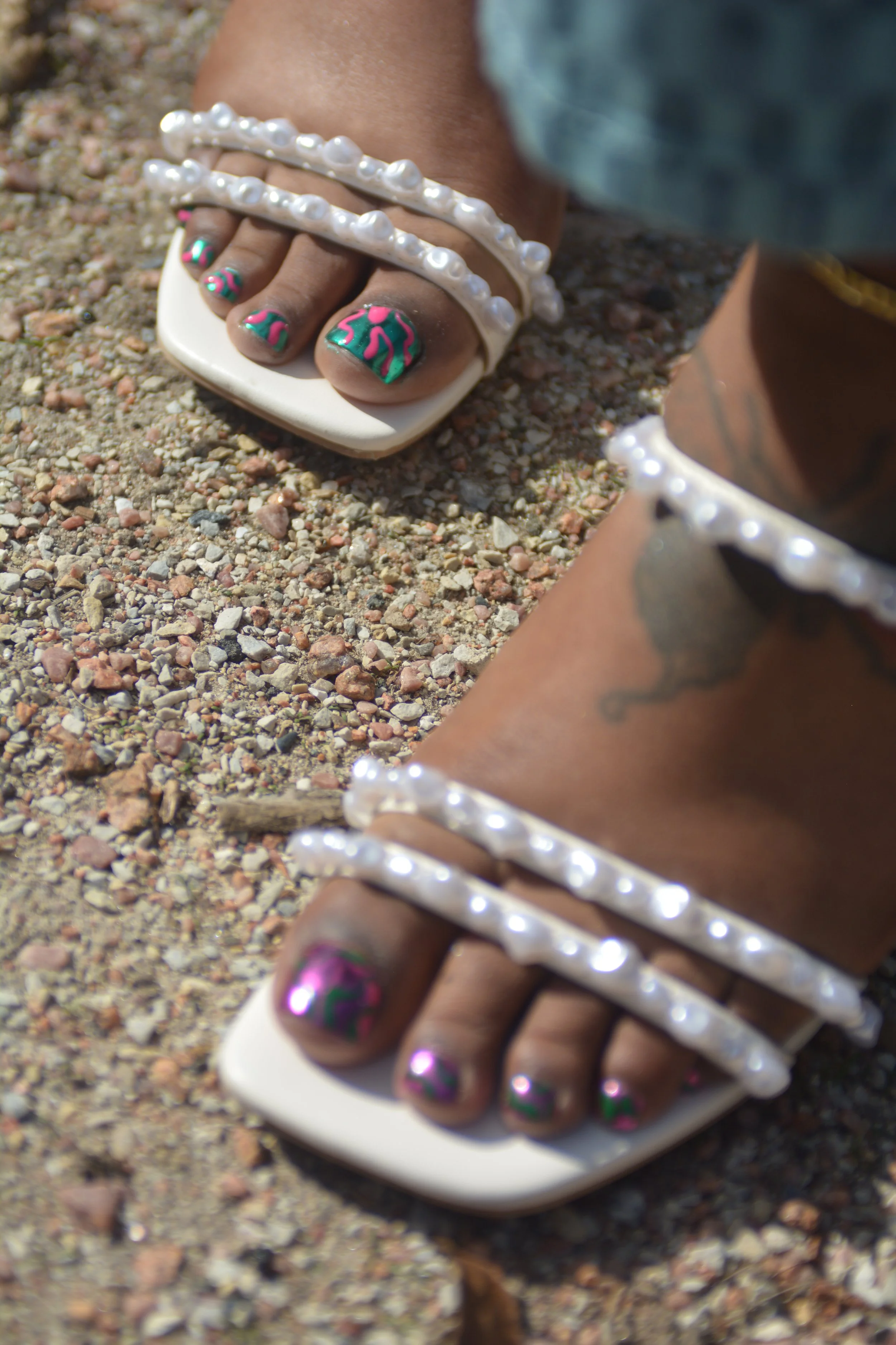 Close-up of a person's toes with colorful nail art, wearing white sandals with pearl embellishments, on a gravel surface. The person also has a pearl anklet and a tattoo on their ankle.