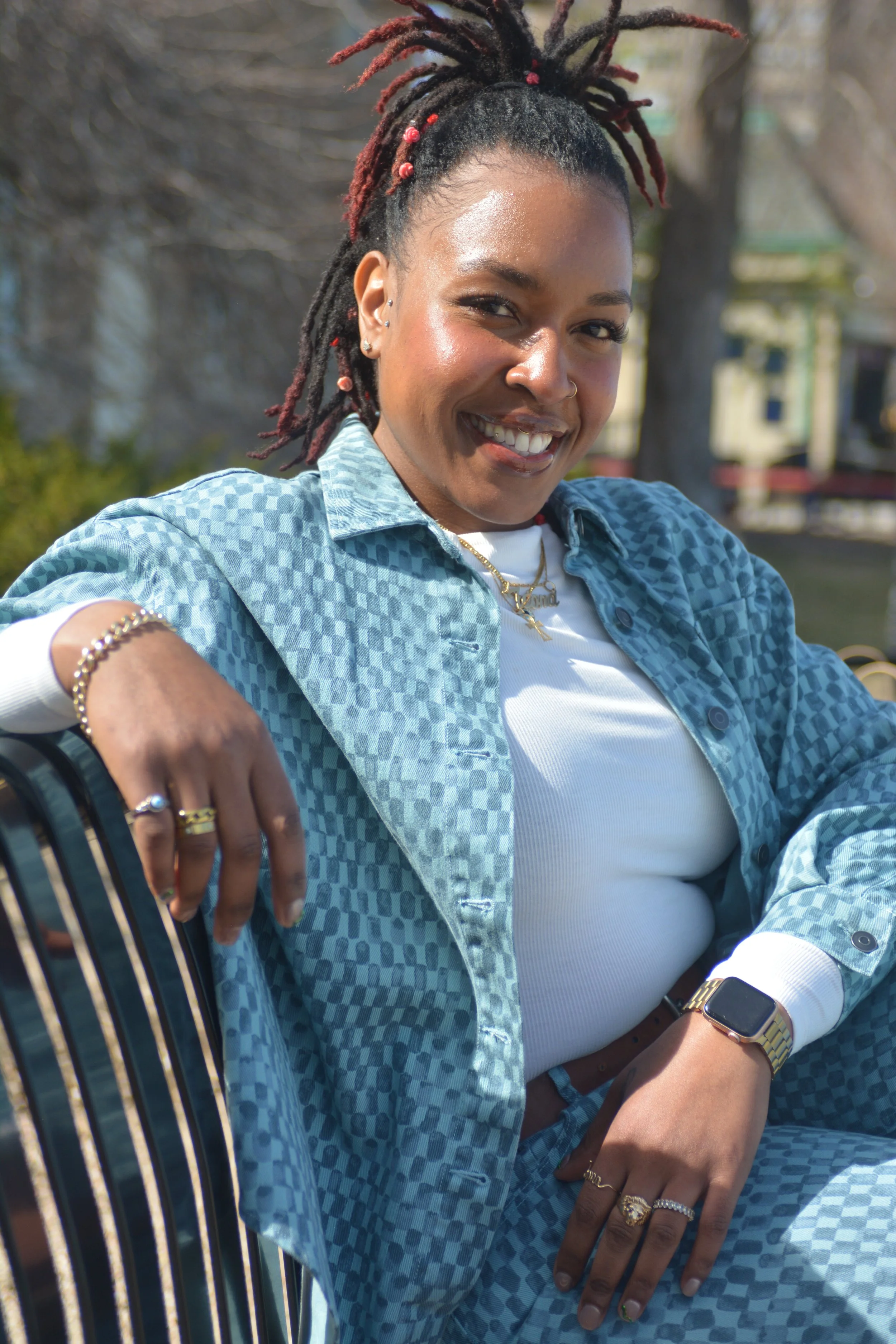 A smiling woman with dreadlocks accented with red beads, wearing a blue checkered shirt over a white top, accessorized with rings, a bracelet, a watch, and layered necklaces, sitting outdoors on a sunny day with blurred trees and a building in the background.