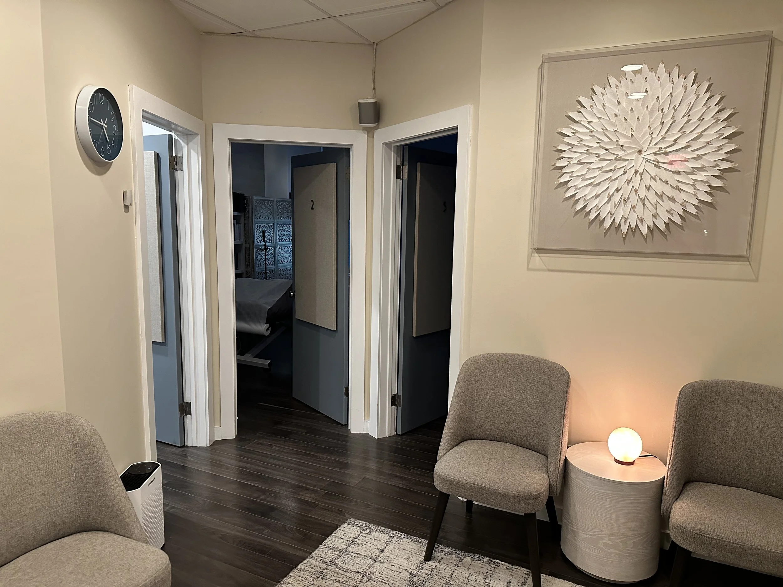 Interior view of a waiting area with two beige chairs, a small round table with a glowing lamp, a wall art piece resembling a white paper flower, and open doors leading to rooms numbered 2 and 3.