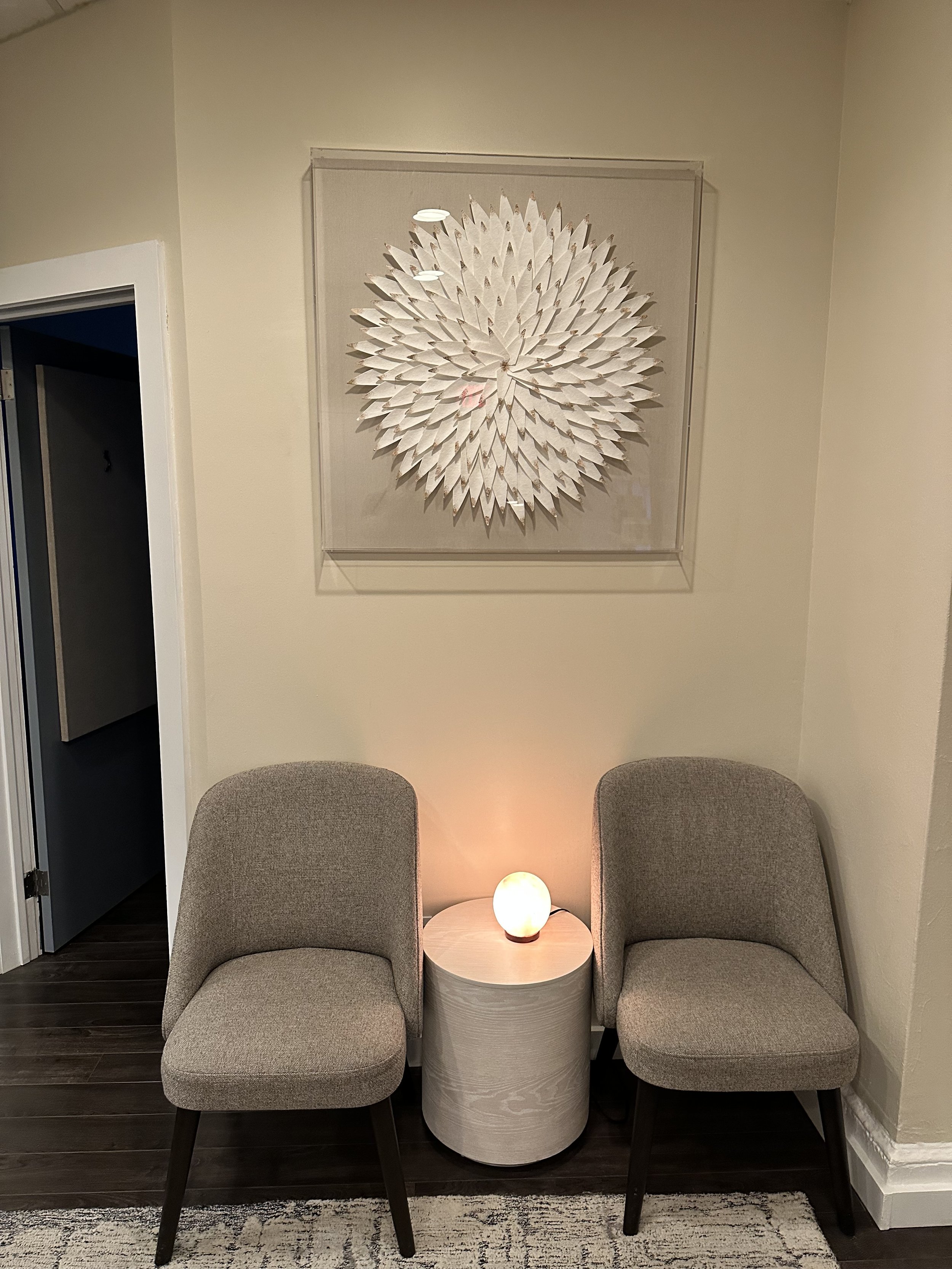 Two beige upholstered chairs with dark wooden legs positioned on either side of a small round wooden table, a glowing orb-shaped lamp on the table, and a decorative wall piece resembling a large flower or starburst located above the chairs.
