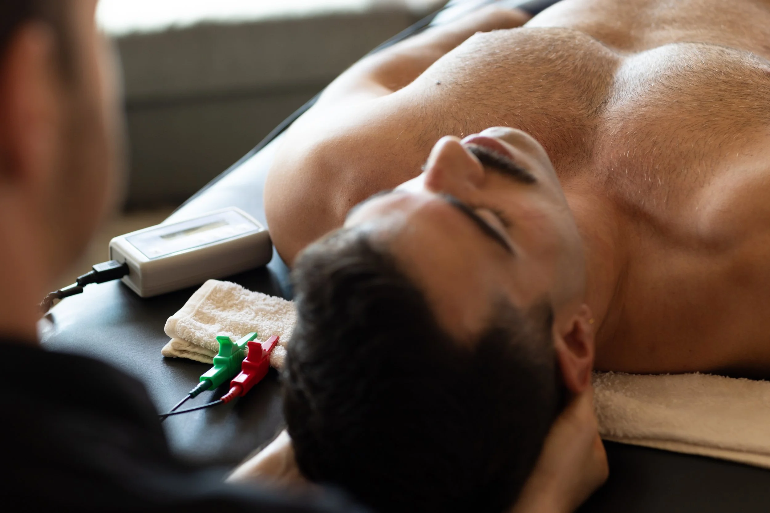 A man lying shirtless on a massage table with electrodes attached to his chest, during a medical procedure or therapy.