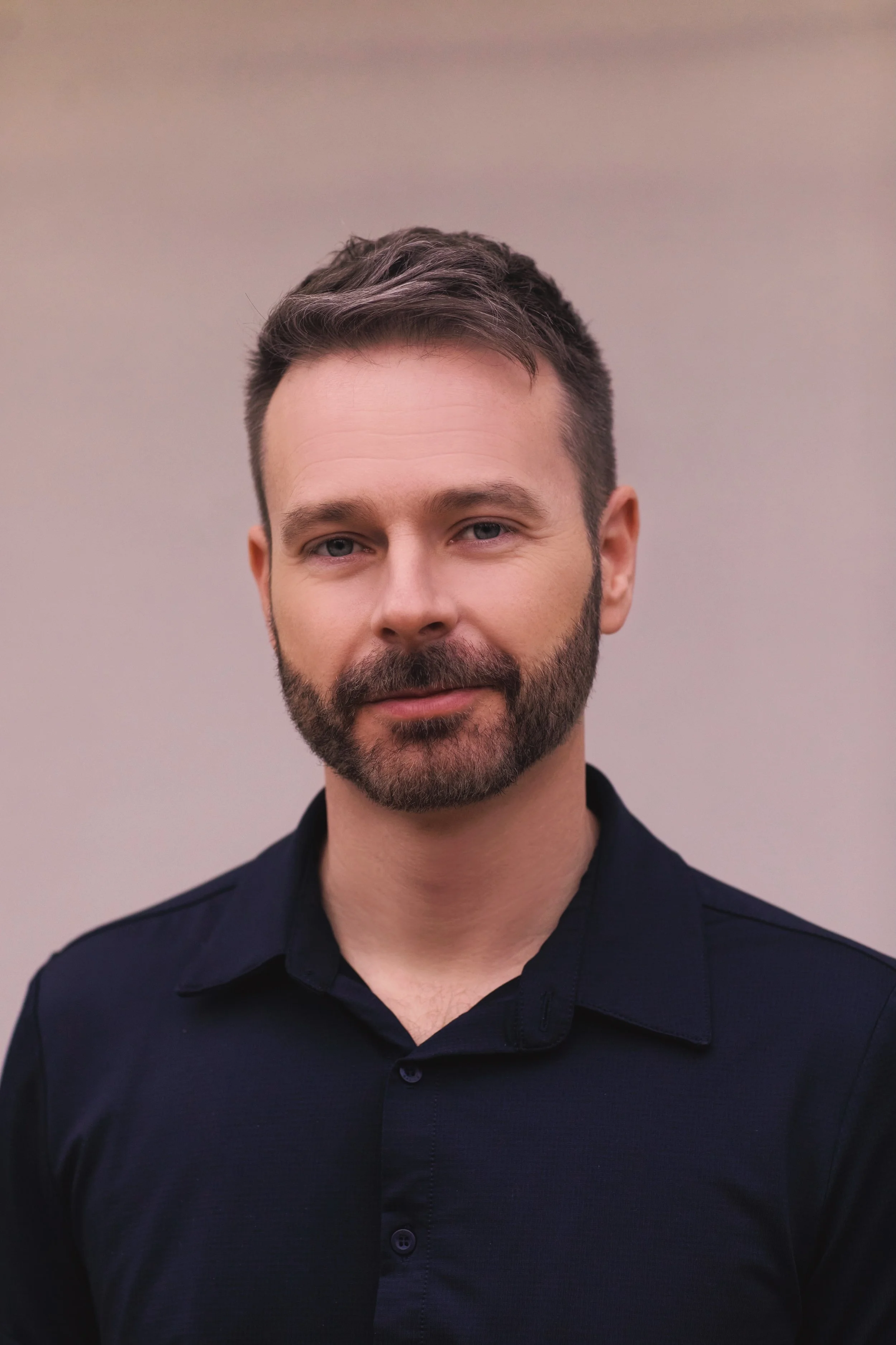 A man with short brown hair, a full beard, and blue eyes wearing a dark shirt, standing against a plain light-colored background.