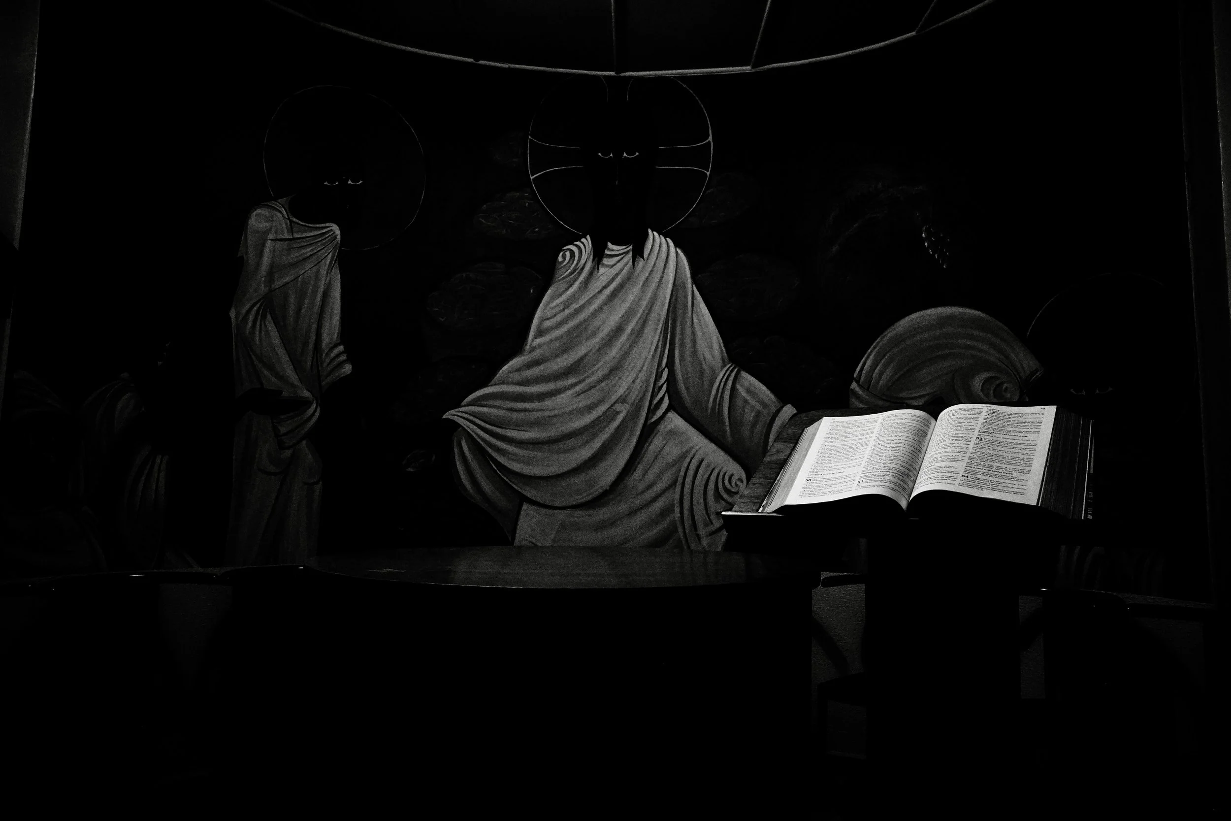 In the quiet of our university chapel, light and shadow intertwine in silent dialogue.
An open Bible rests in the foreground — a symbol of wisdom, reflection, and the search for meaning — before a figure that embodies serenity and compassion.
It is a