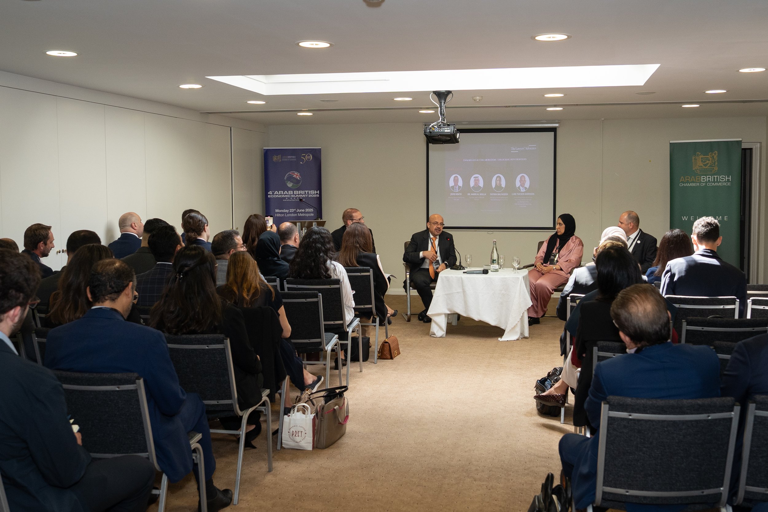 A professional conference with a panel of three speakers at a round table in front, addressing an audience seated in rows. The room has a projector screen displaying a presentation and banners for the Arab British Chamber of Commerce and Arab British