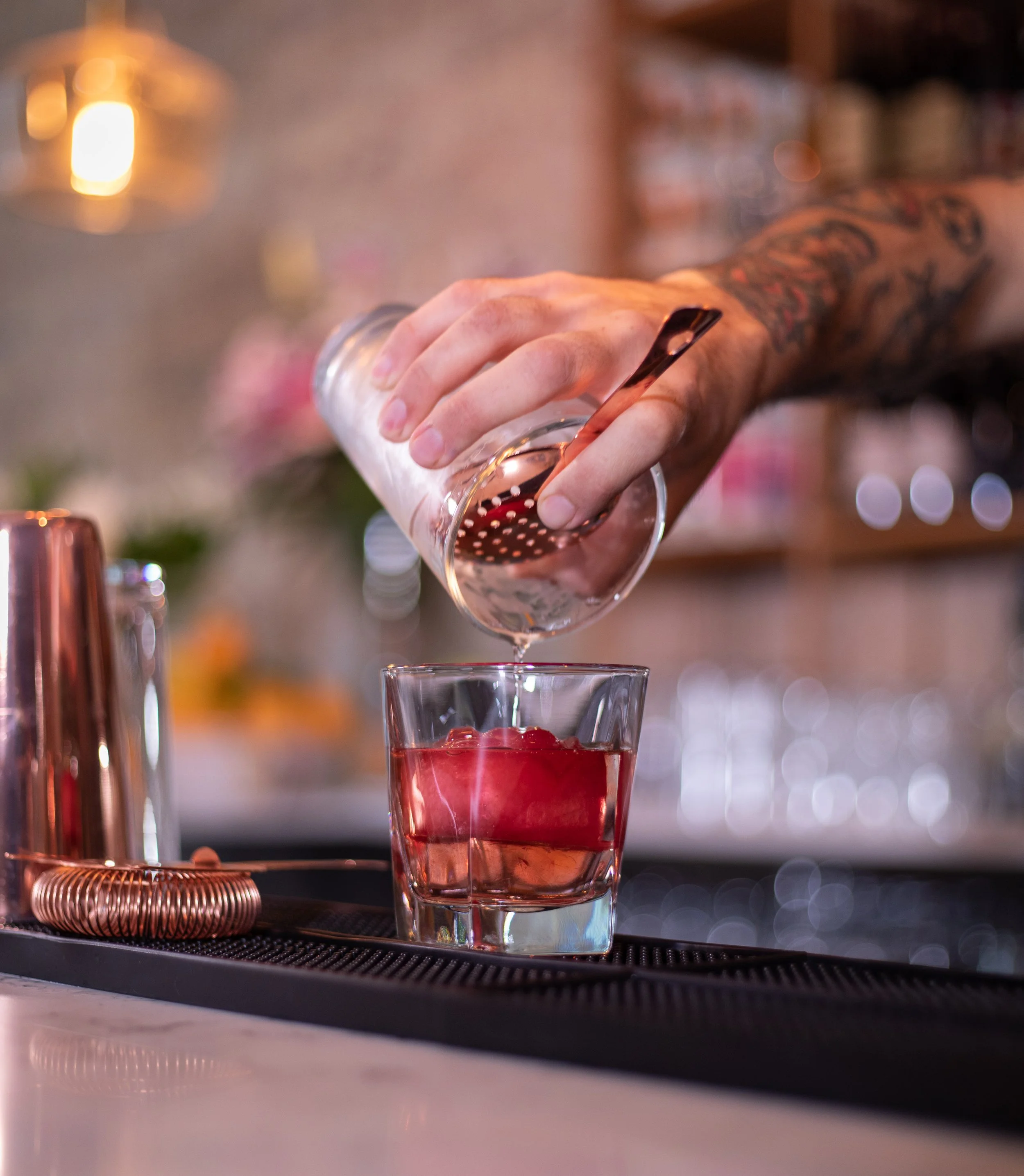 Bartender pouring clear liquid through a strainer into a glass with ice and red fruit at a bar.