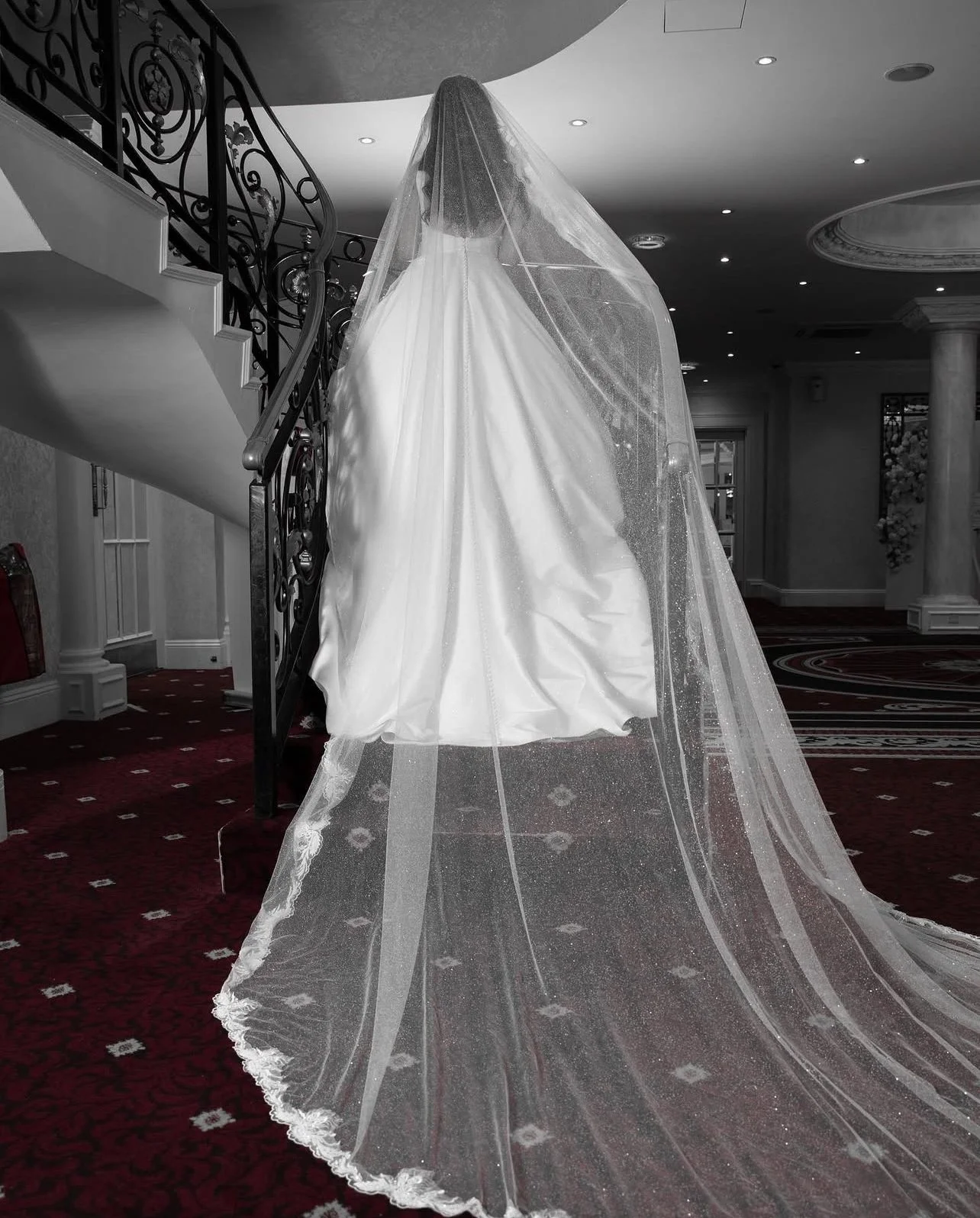 A white wedding dress with a long train and veil hanging over a staircase inside a building with ornate architecture.