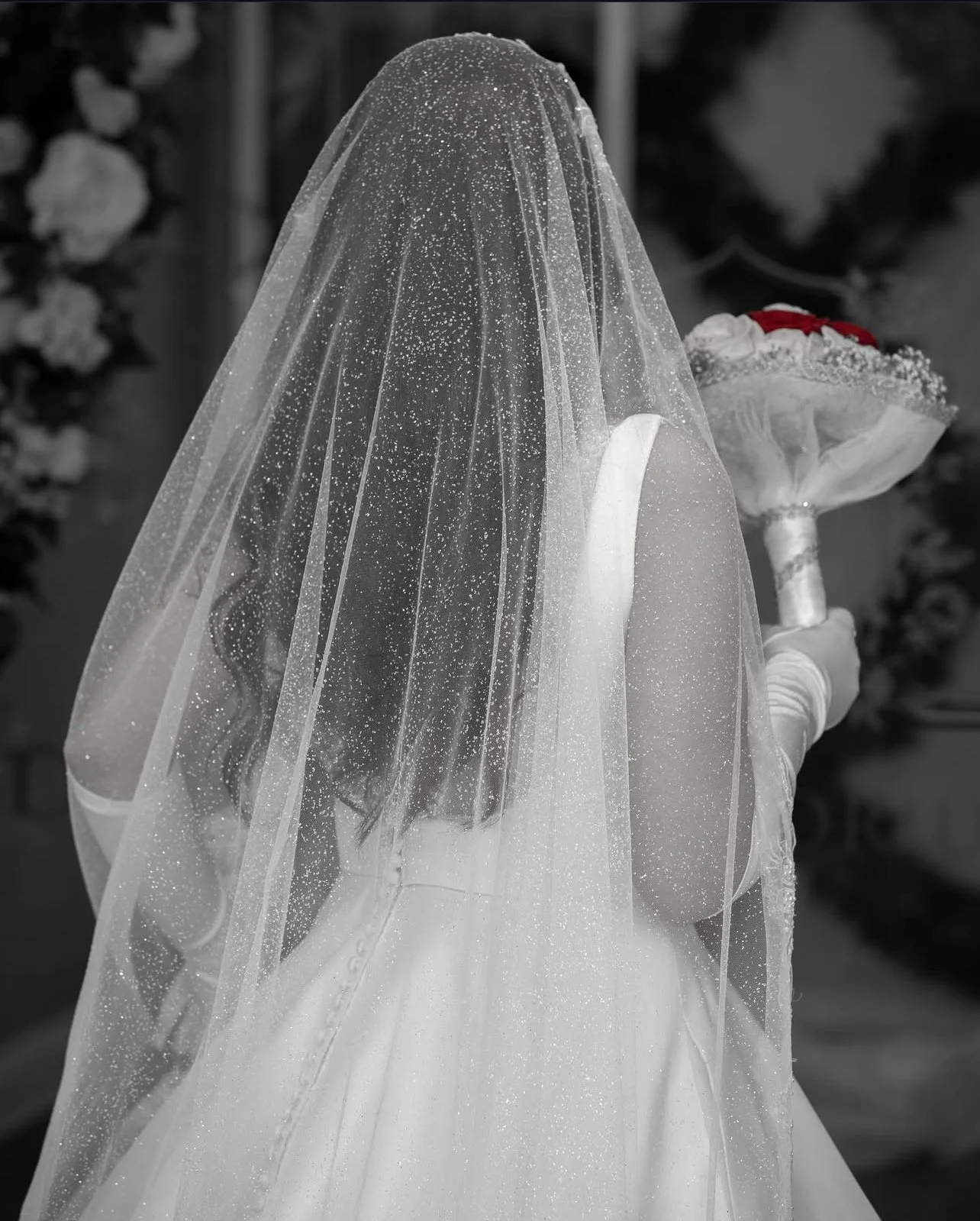 A bride standing with her back, wearing a wedding dress and a veil covered with water droplets, holding a bouquet of flowers.