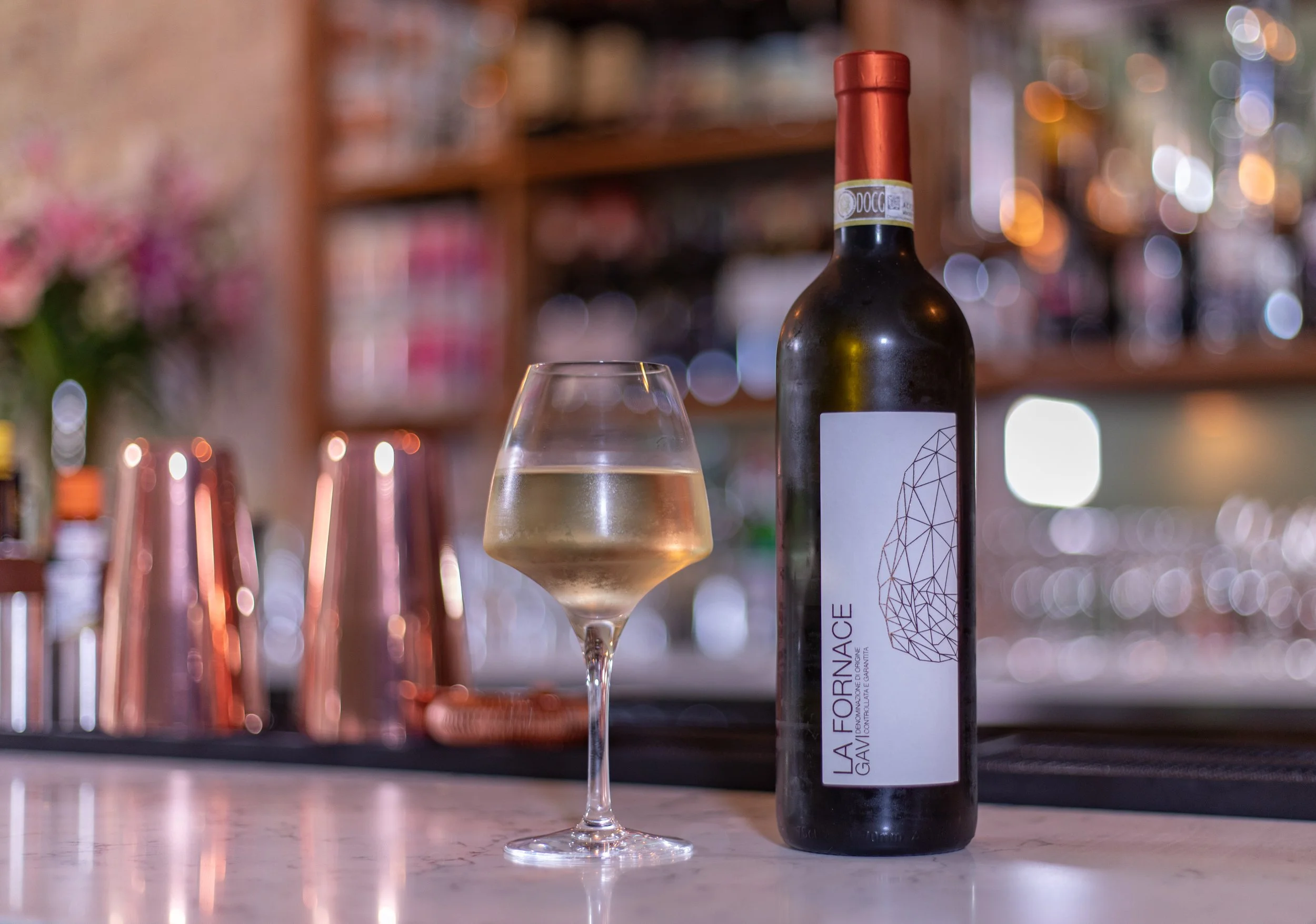 A bottle of La Fornace wine and a glass of white wine on a bar counter with a blurred bar background and pink flowers