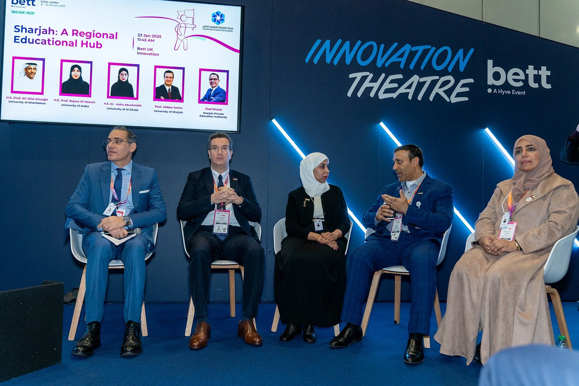 Five individuals seated on a stage during a panel discussion at the Innovation Theatre event. They are dressed in business attire and wearing conference badges. The background features a large screen displaying information about the event titled 'Sha
