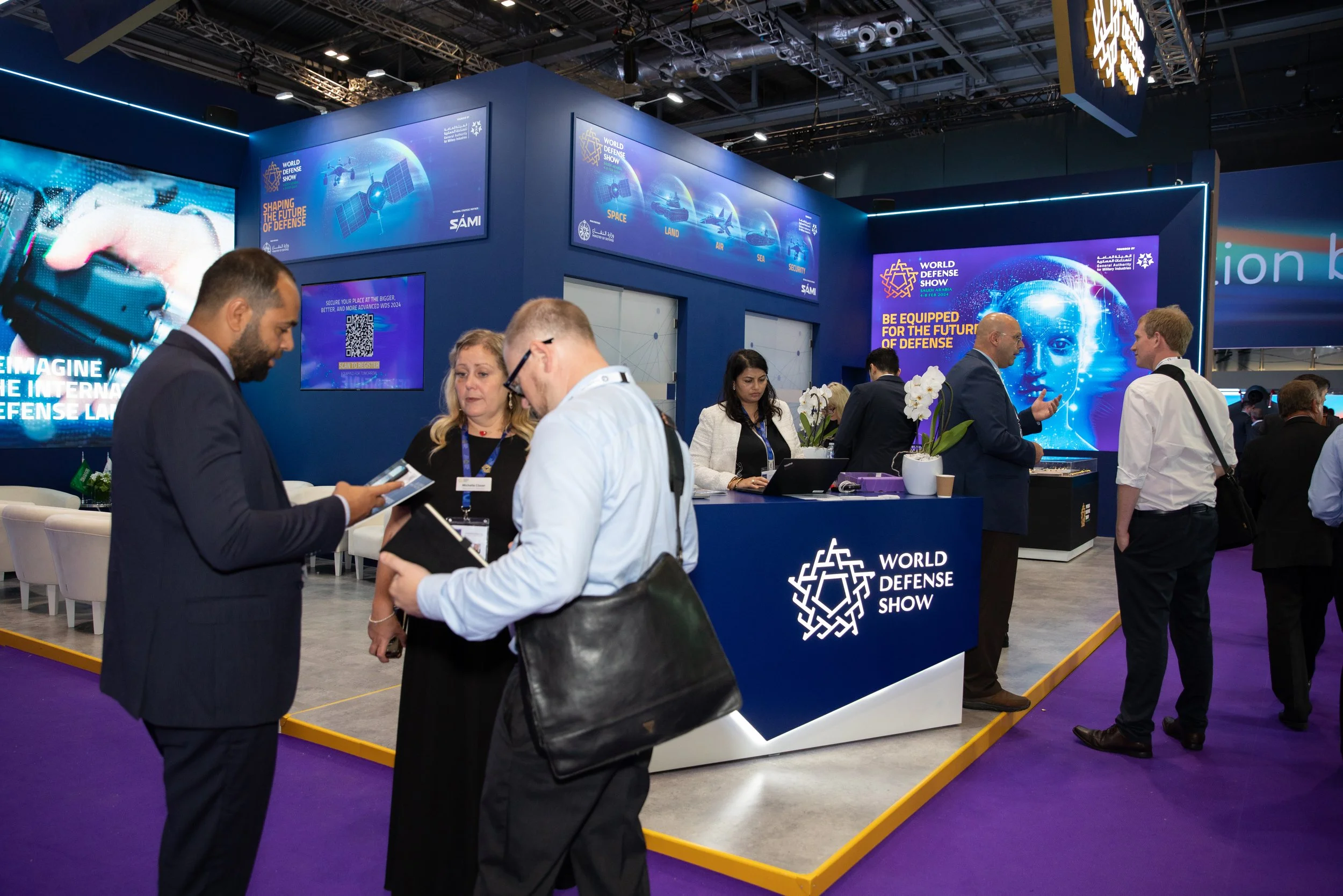 People at the World Defense Show booth, engaging in conversations and managing displays in a large convention hall with digital screens and promotional materials about defense technologies.