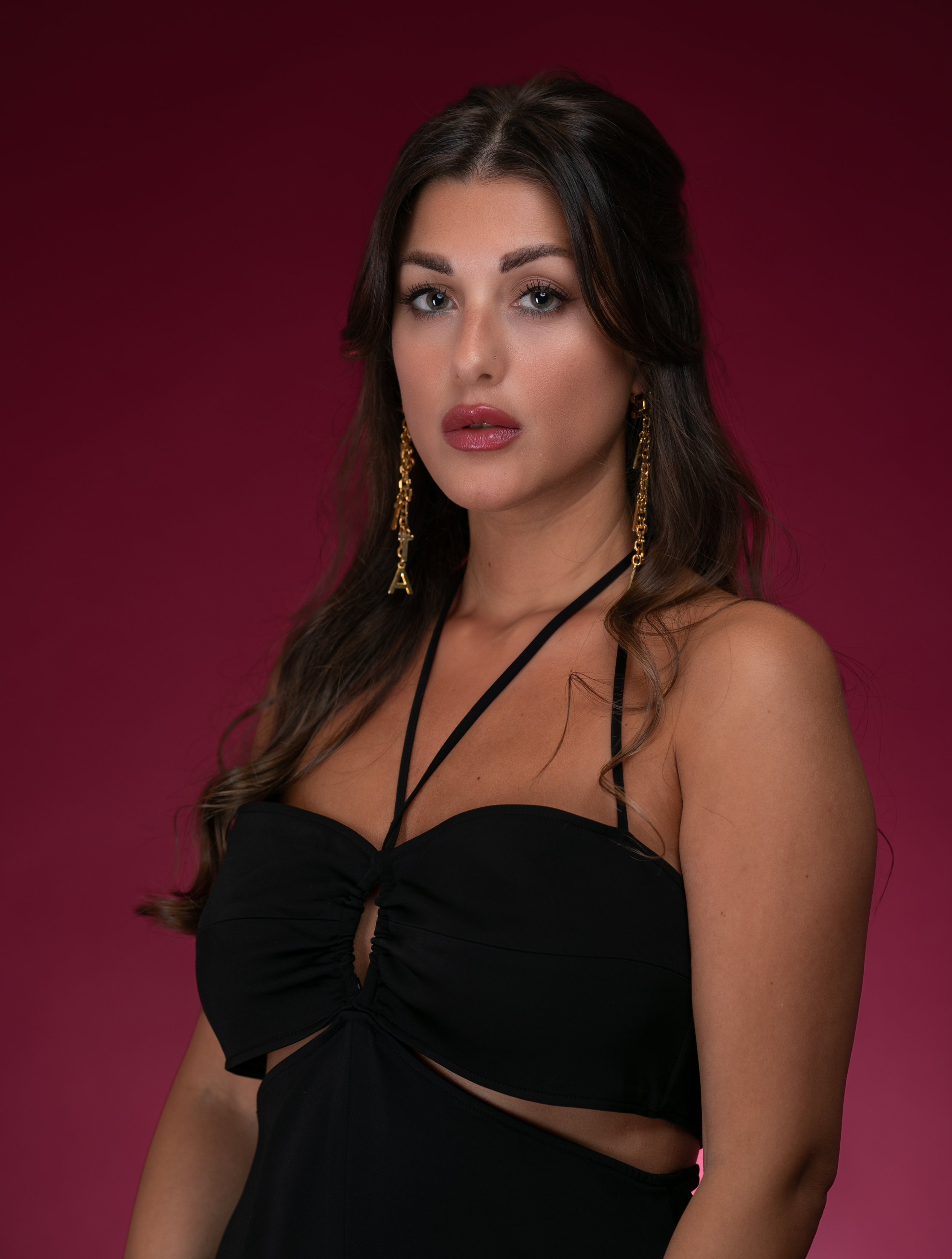 A woman with wavy dark brown hair, wearing gold earrings, a black strap dress with cutouts, posed against a dark pink background.