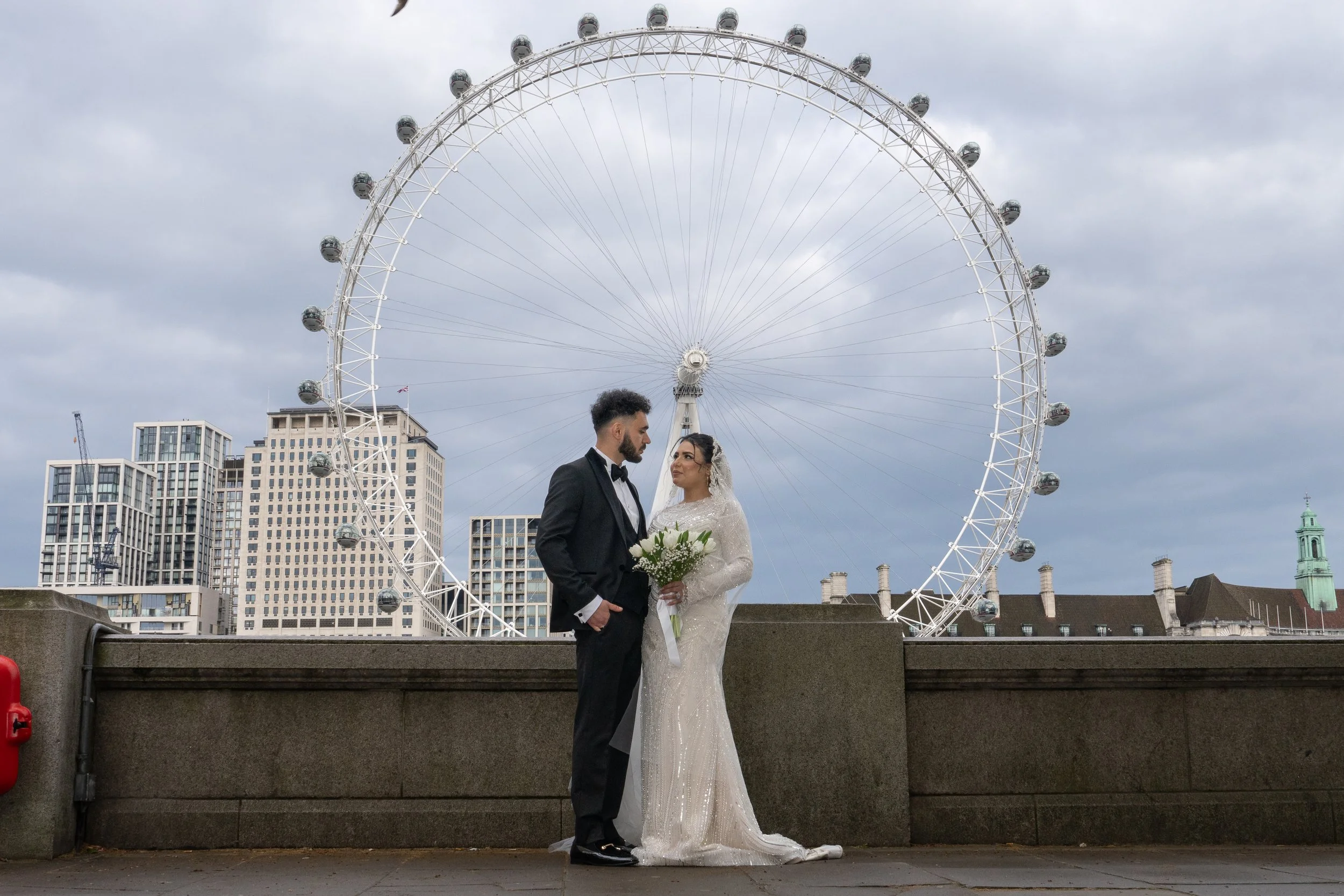 A bride and groom in wedding attire standing on a city waterfront with a Ferris wheel and tall buildings in the background.