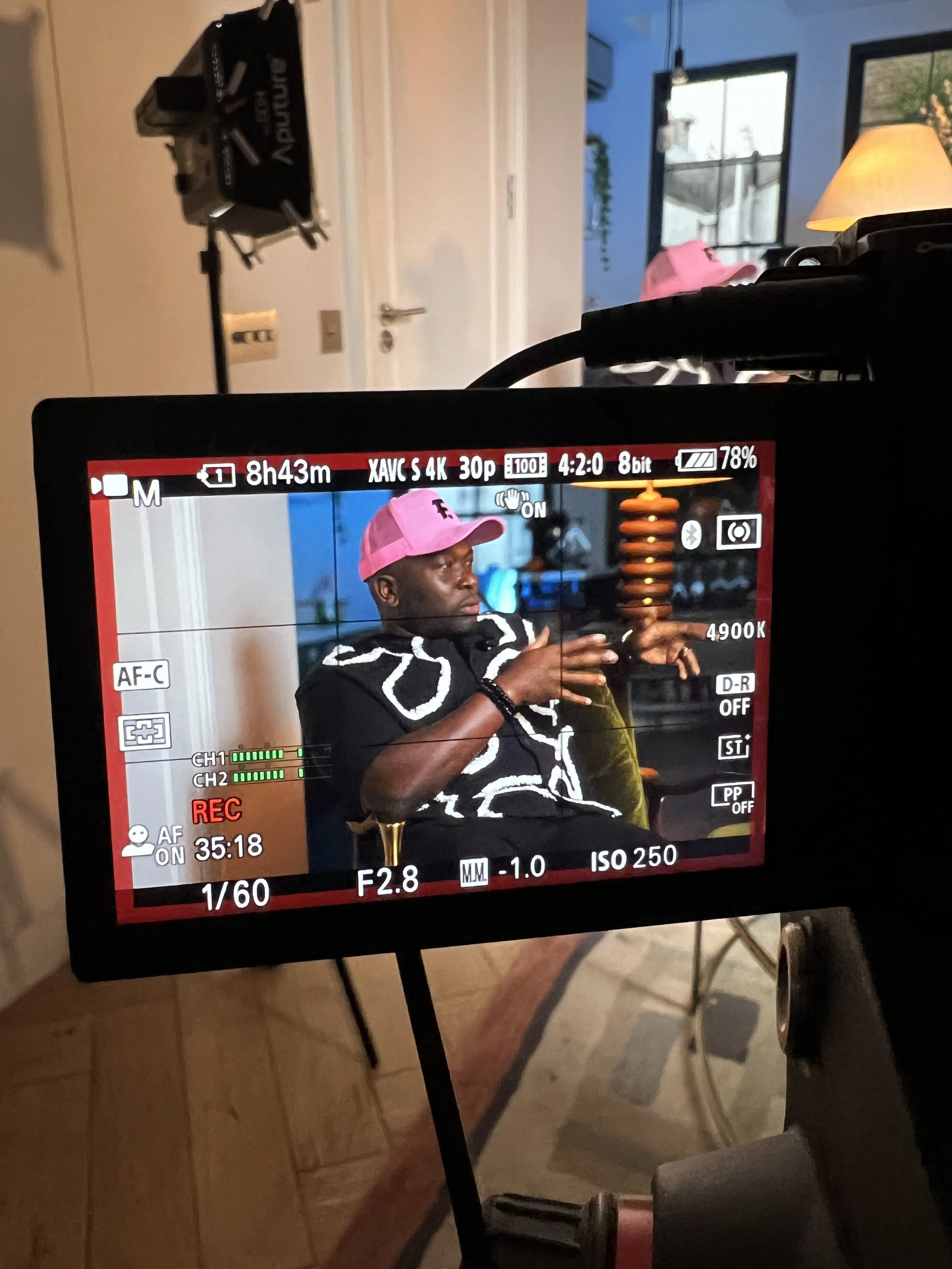 Camera screen showing a person in a pink cap and black shirt with white designs, sitting on a yellow chair inside a room with large windows and a lamp, as part of a video recording setup.