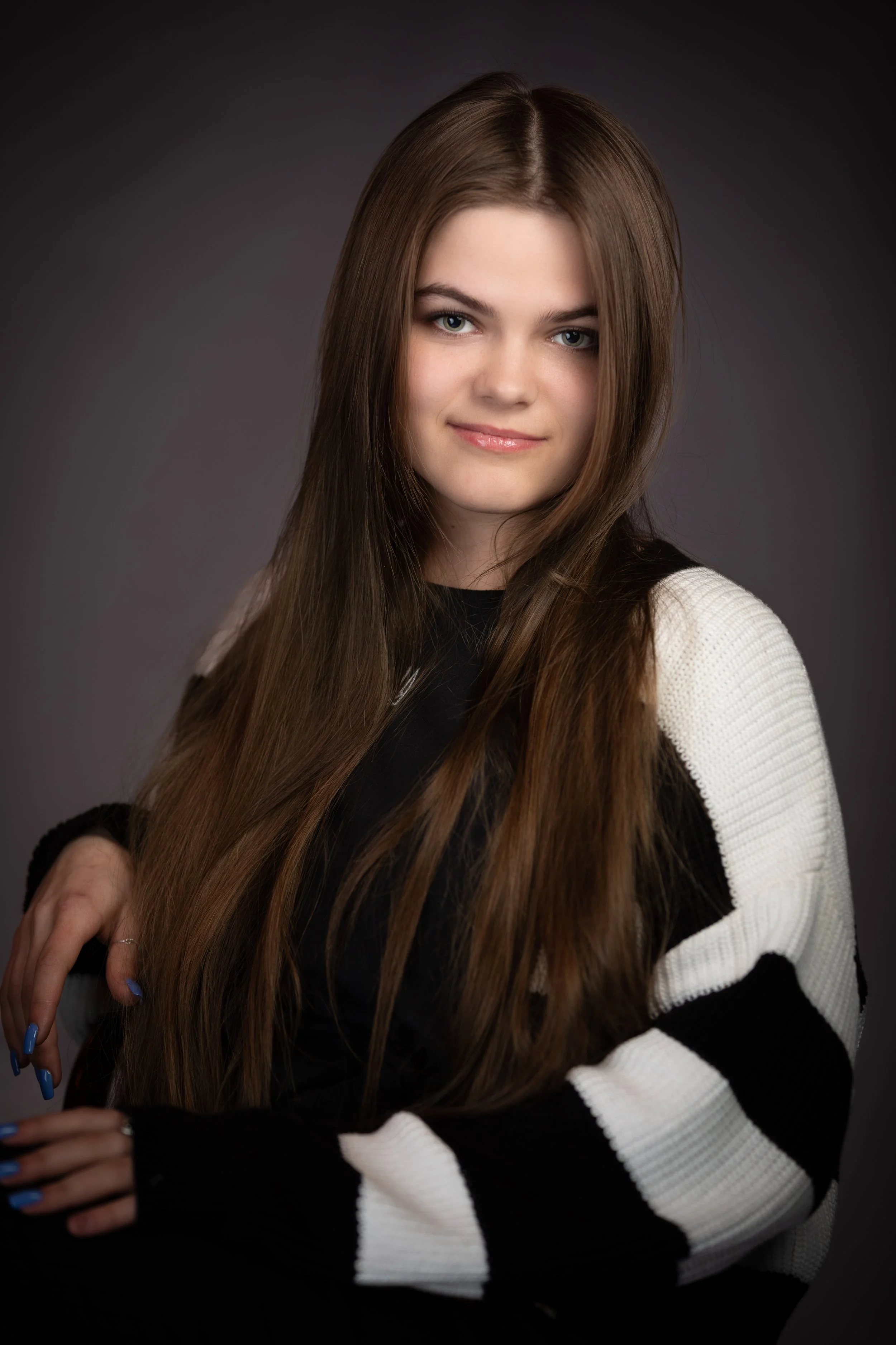 Young woman with long brown hair, wearing a black and white sweater, looking at the camera with a slight smile, against a dark gray background.
