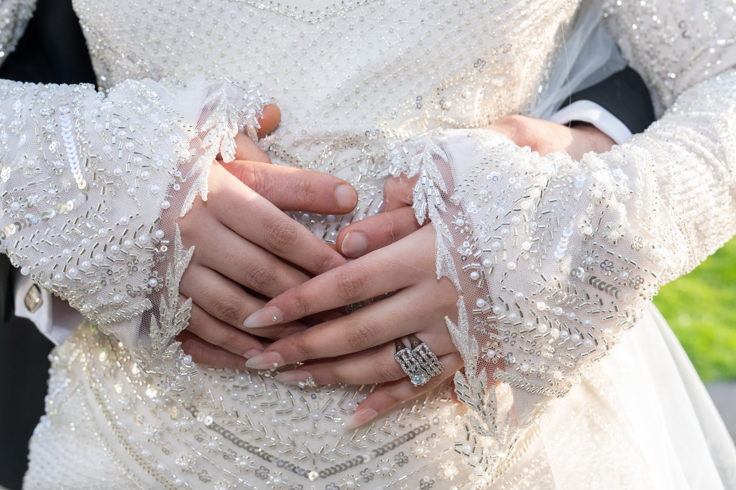 Close-up of a couple’s hands with wedding rings, gently touching each other, on a bride in a detailed, beaded wedding gown with long sleeves.