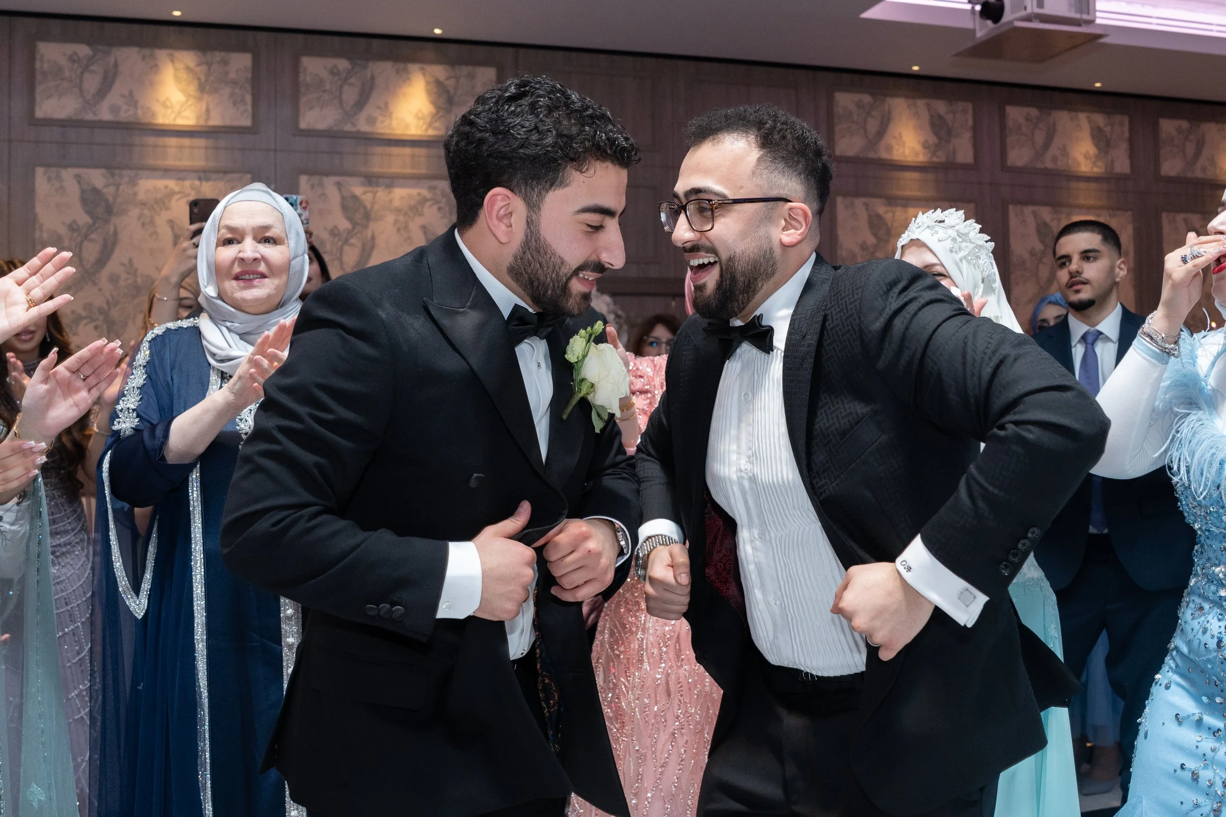 Two men in tuxedos dancing and smiling at a wedding reception, surrounded by guests in elegant attire, including women in colorful dresses and headscarves.