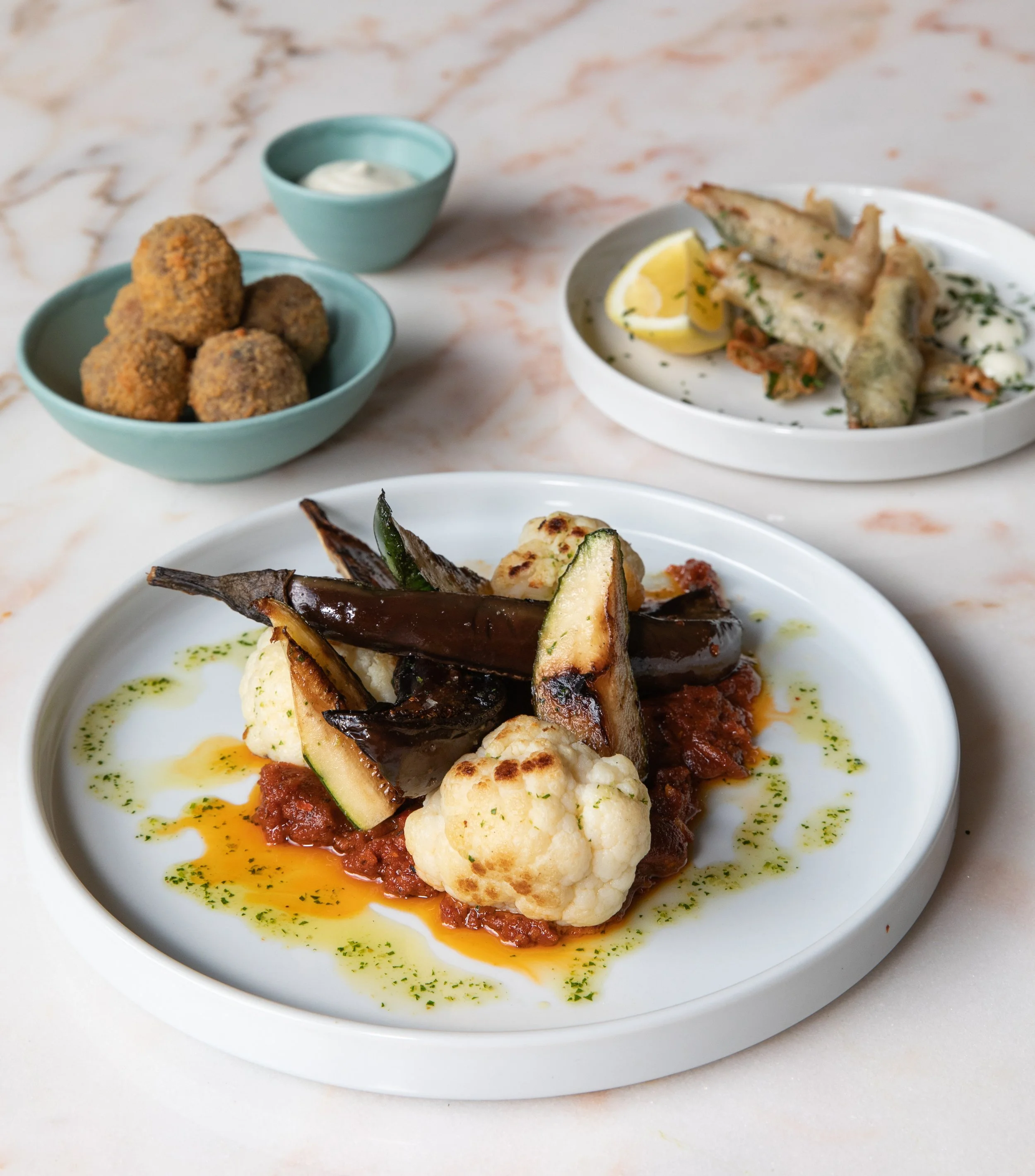 A plate of roasted vegetables including cauliflower, zucchini, and eggplant, served with tomato sauce and garnished with herbs. In the background, there are other plates with fried fish and fried vegetables, and a bowl of croquettes with a small dish