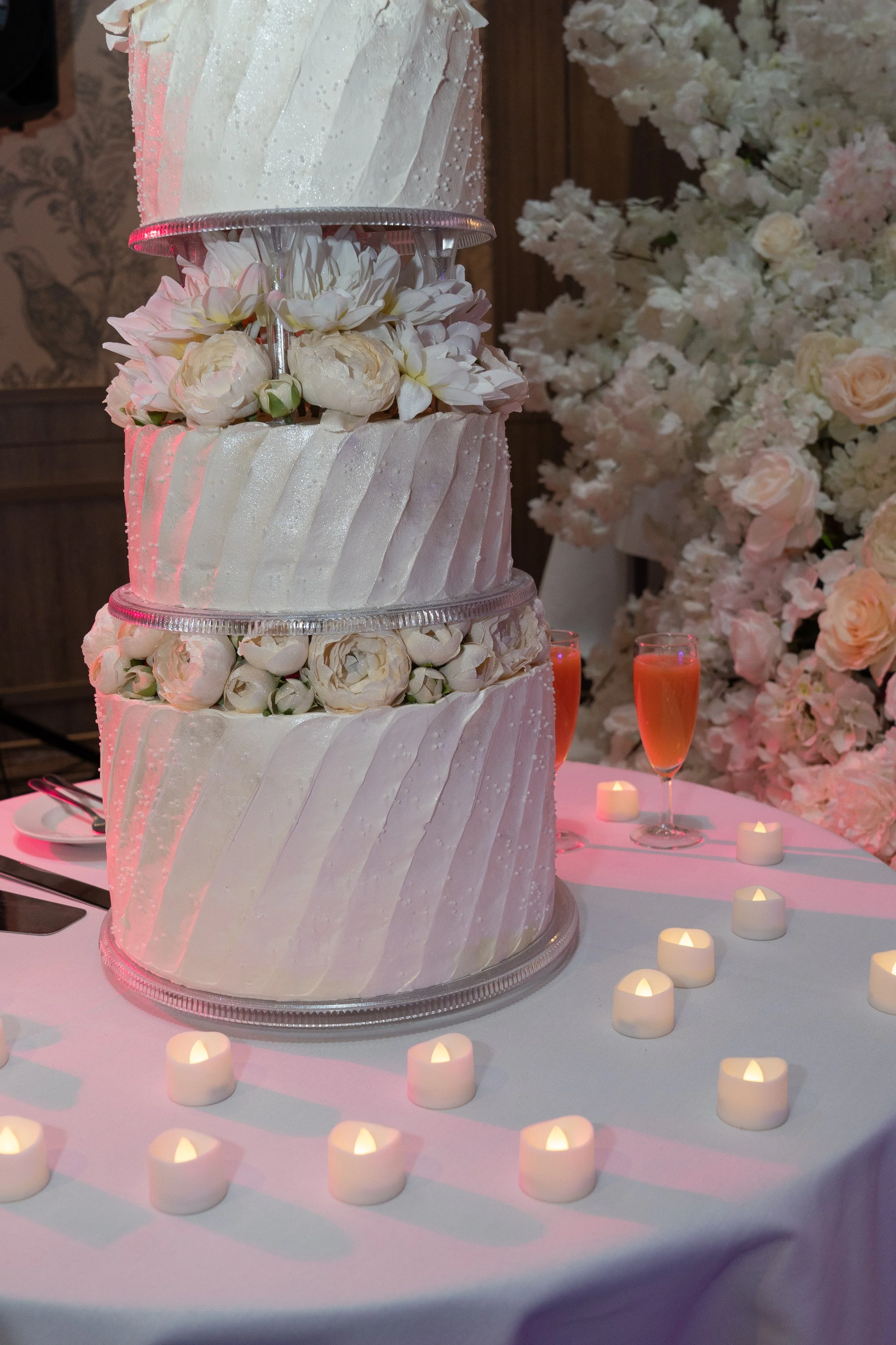 A four-tiered wedding cake decorated with white icing, white roses, and peonies, surrounded by small candles on a table with a white tablecloth, pink drinks, and a large floral arrangement in the background.