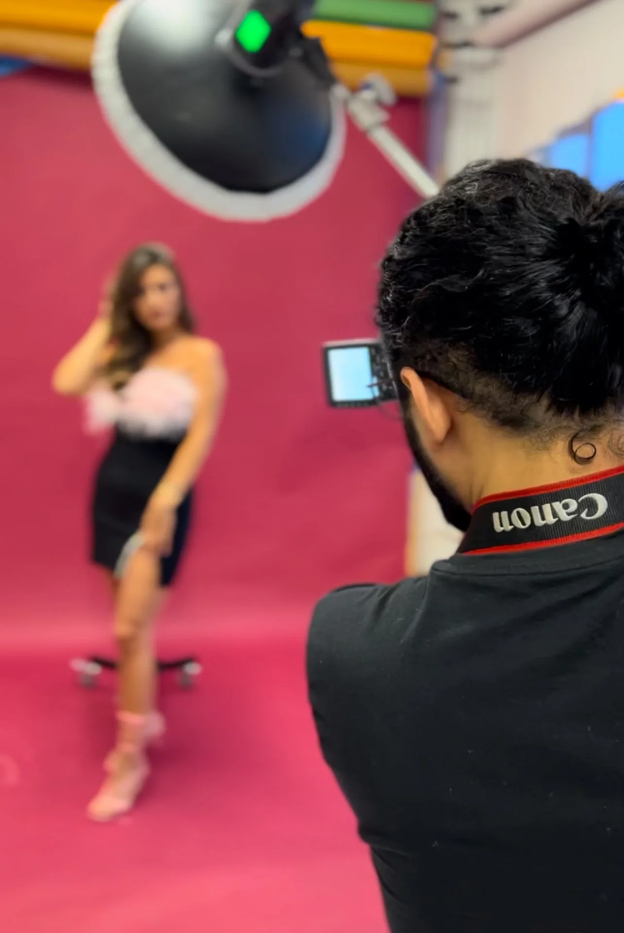 A photographer is taking a picture of a woman posing in front of a pink backdrop. The photographer's back is visible, with a camera strap reading "Canon". The woman is dressed in a stylish, strapless dress and high heels, standing with her hand on he