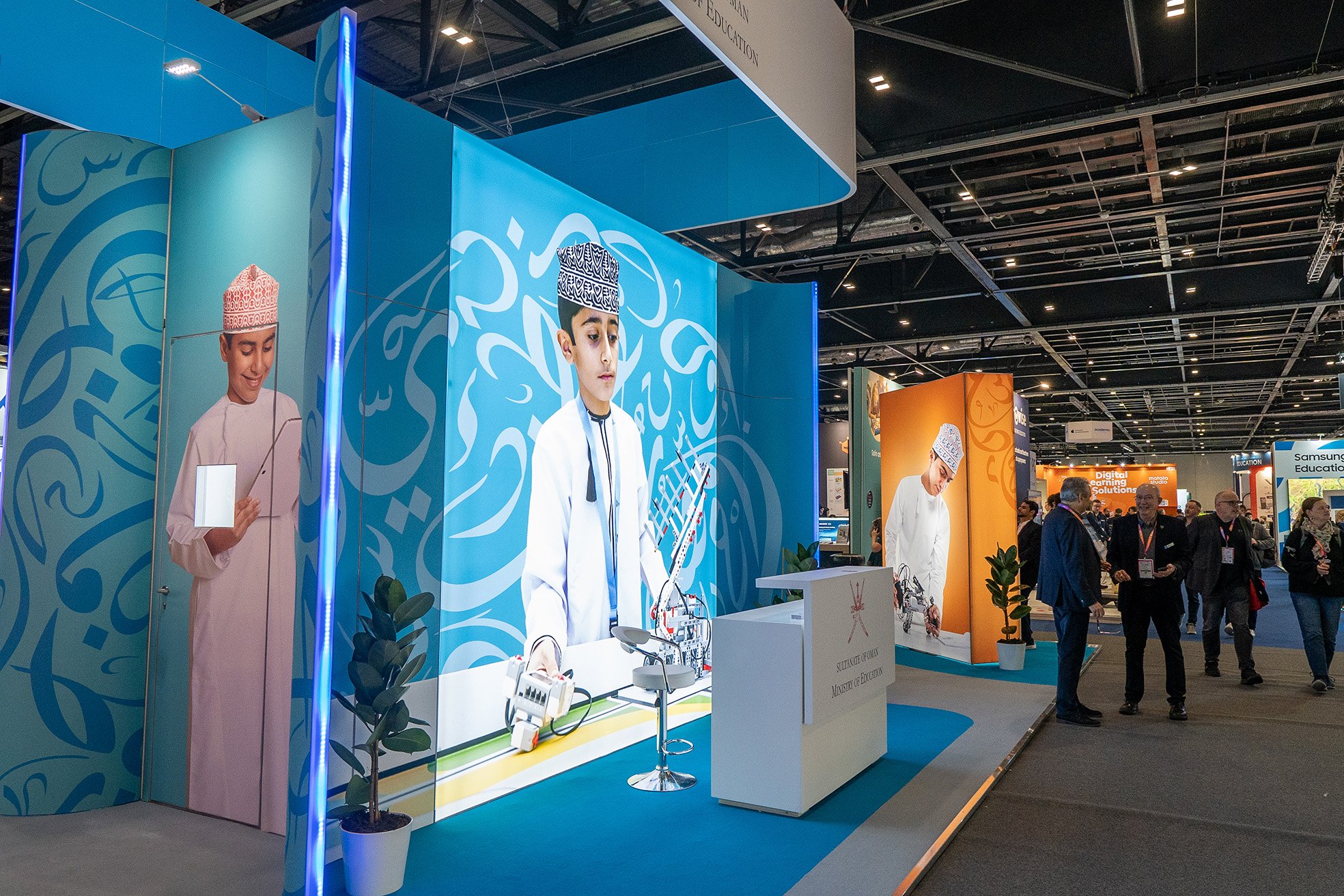 A booth at an exhibition with large images of young boys in traditional Omani clothing and a model robot on display. Visitors are walking and talking nearby.