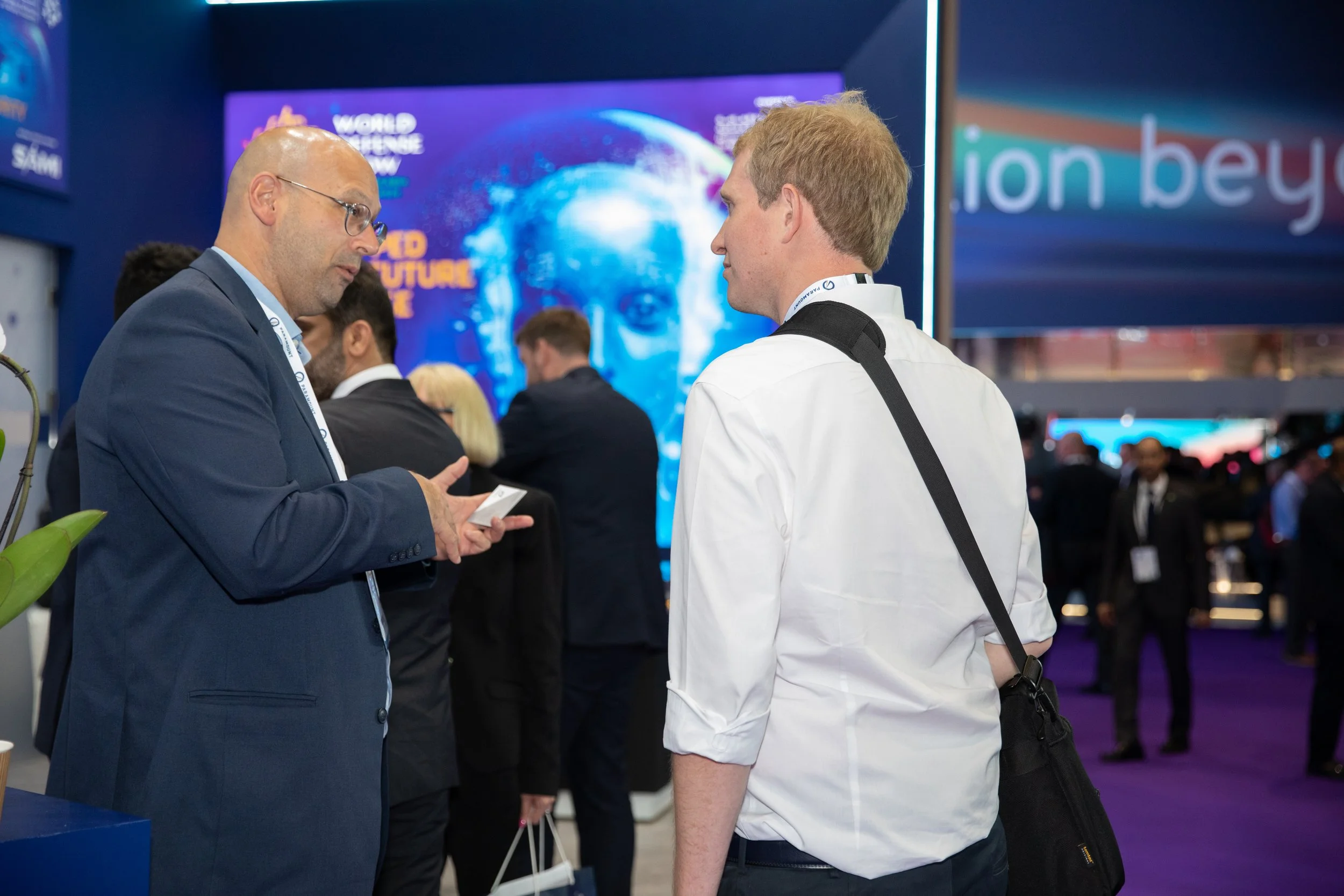 Two men in business attire having a conversation at a conference, with other attendees and a large digital display screen in the background.