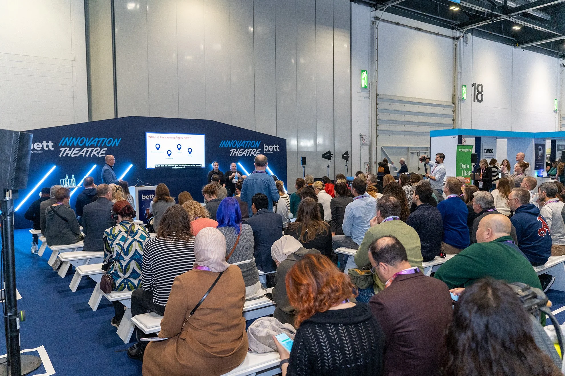 A conference or seminar with a large audience seated and facing a stage where a presenter is speaking. The stage has a large screen displaying information, and the backdrop has the words 'Innovation Theatre' and 'bett'.