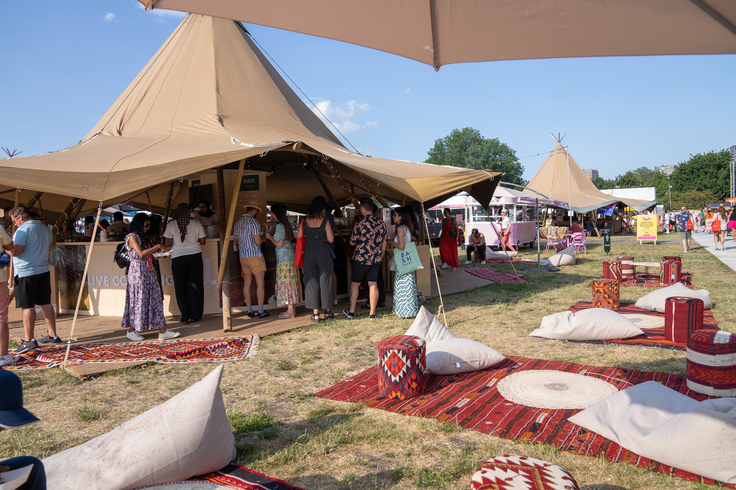 People standing in line at a food or drink stand with tents and outdoor seating at a festival or fair on a sunny day.