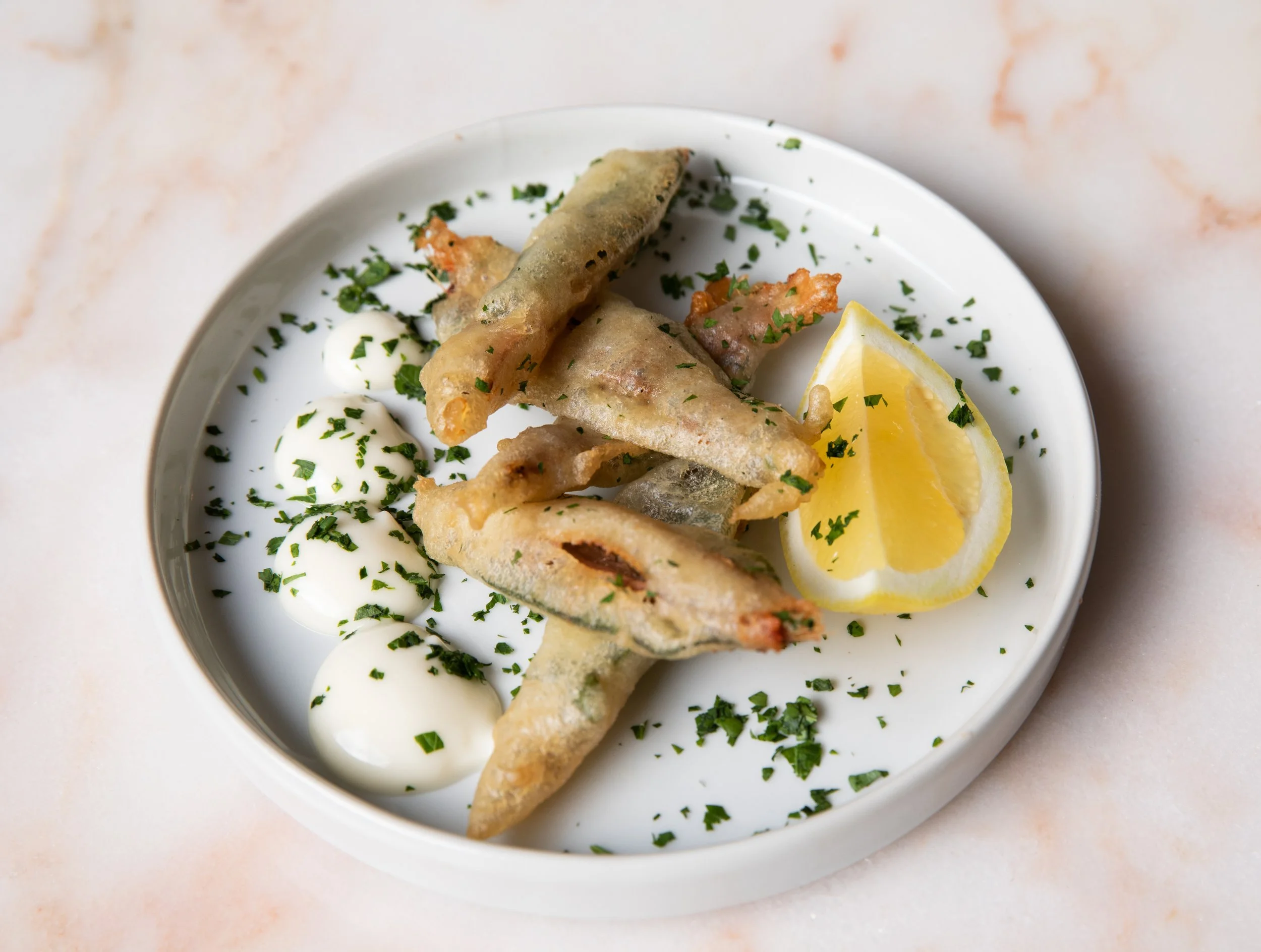 Plate of fried fish fillets with lemon wedge, small dollops of tartar sauce, and chopped parsley garnished on top.