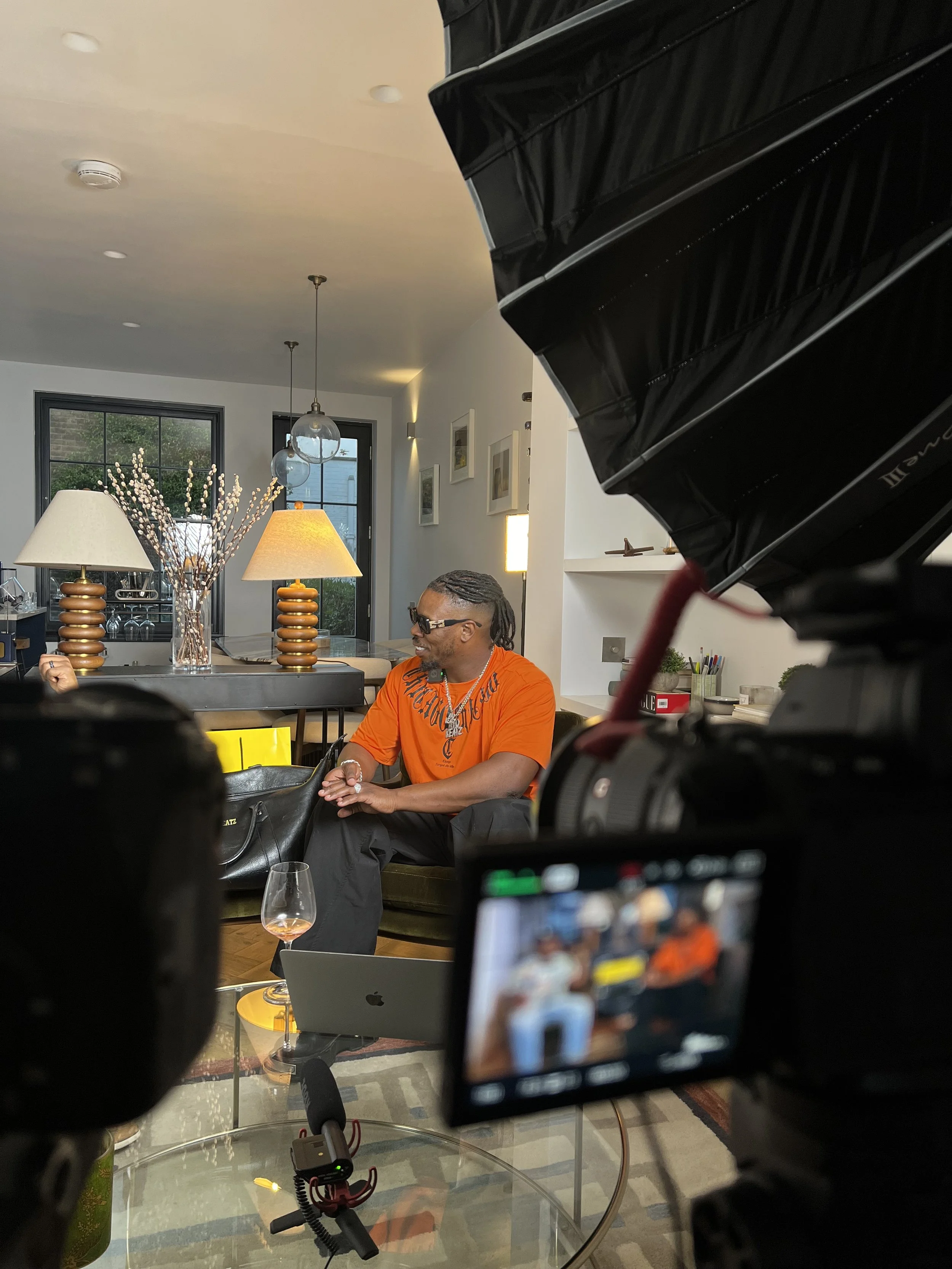 Man with dreadlocks wearing orange shirt and sunglasses sitting on a couch during a video interview or recording, with a recording camera in the foreground and a glass of drink on a table nearby.