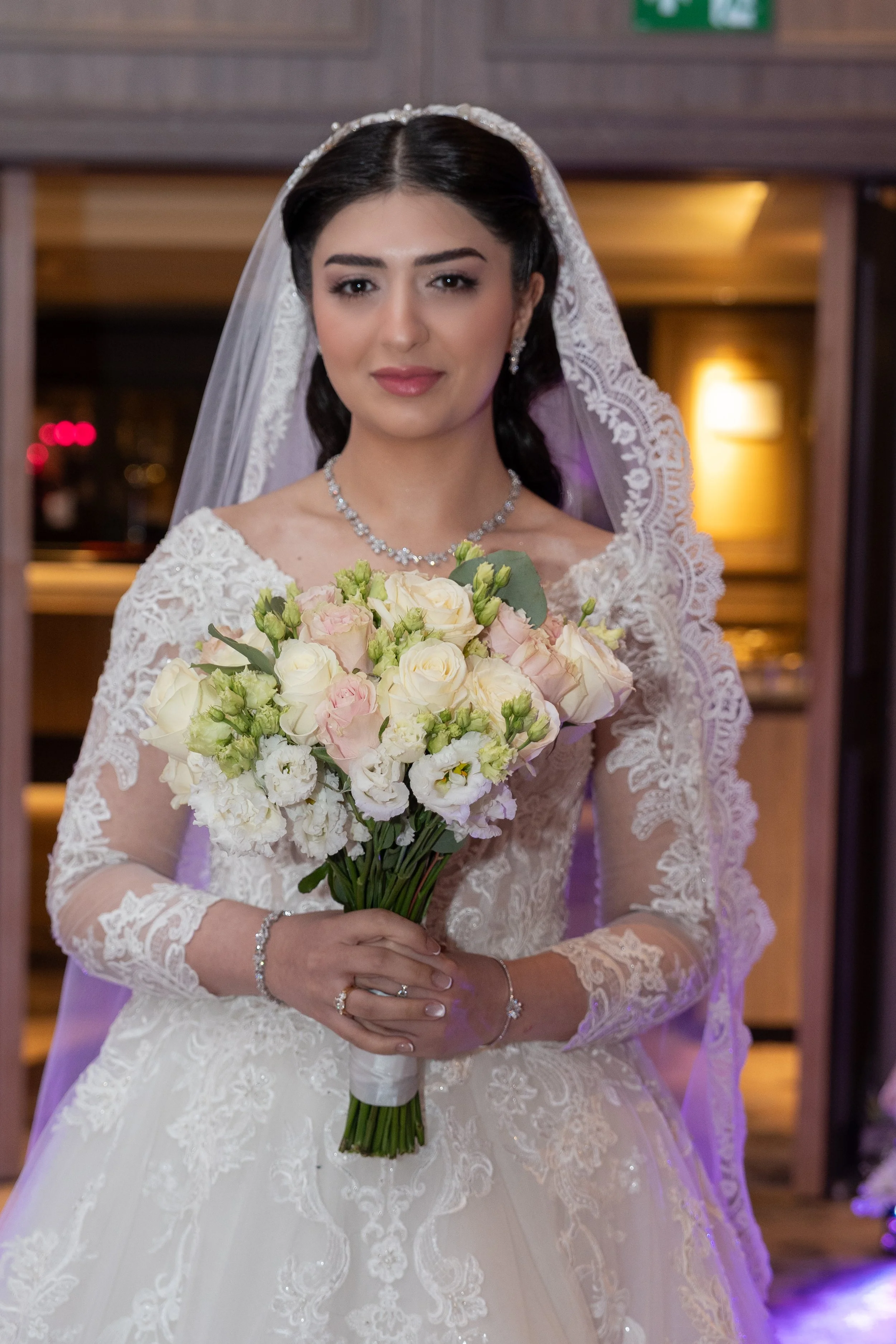 A bride with dark hair, wearing a white lace wedding dress, pearl jewelry, and a lace veil, holding a bouquet of white and light pink flowers.