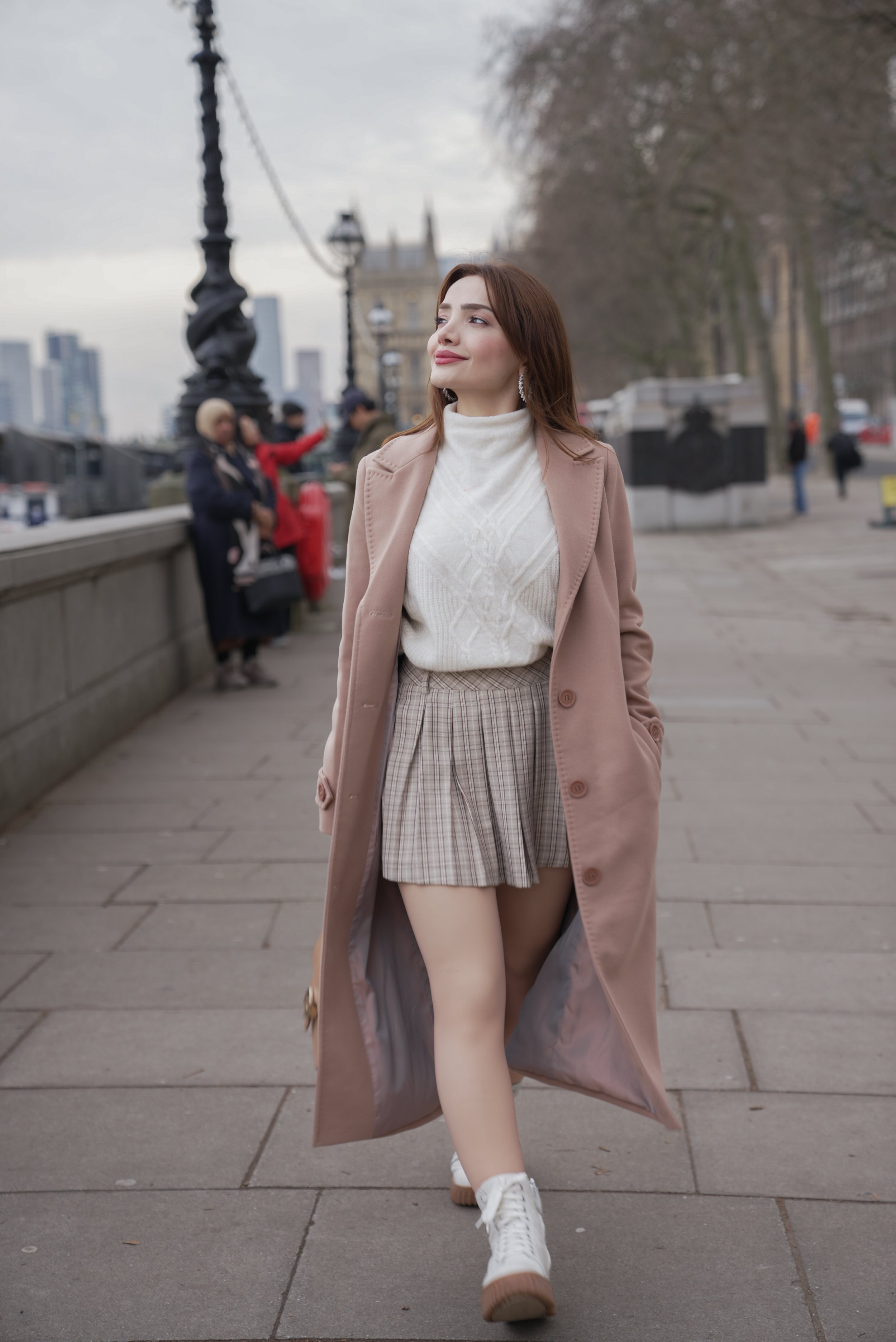 A woman in a beige coat, white sweater, plaid skirt, and white sneakers walking along a city street overcast with trees and iconic London architecture in the background.