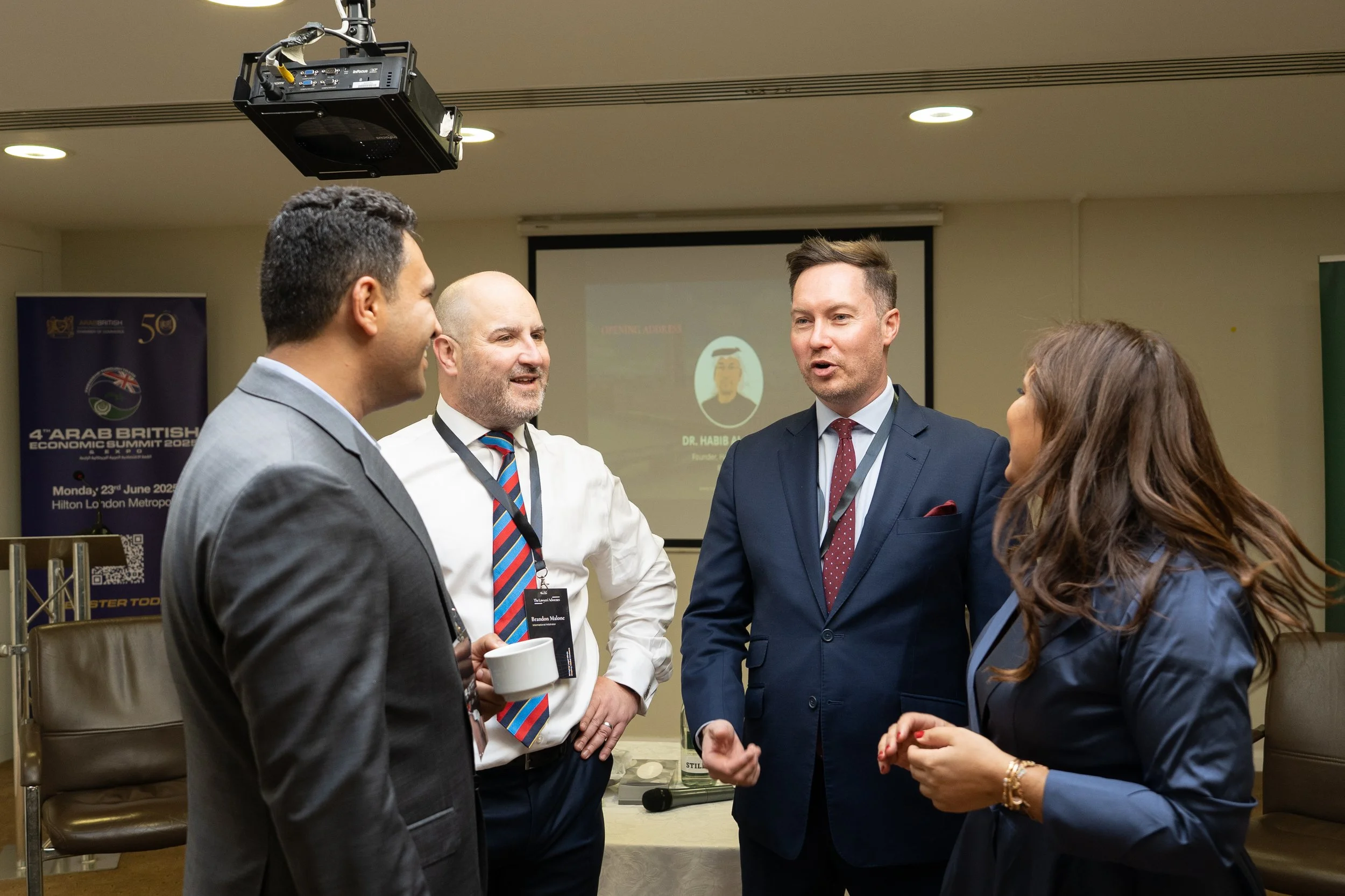 Four business professionals, three men and one woman, engaged in conversation at a conference or meeting in a hotel conference room. A presentation screen and banner are visible in the background.