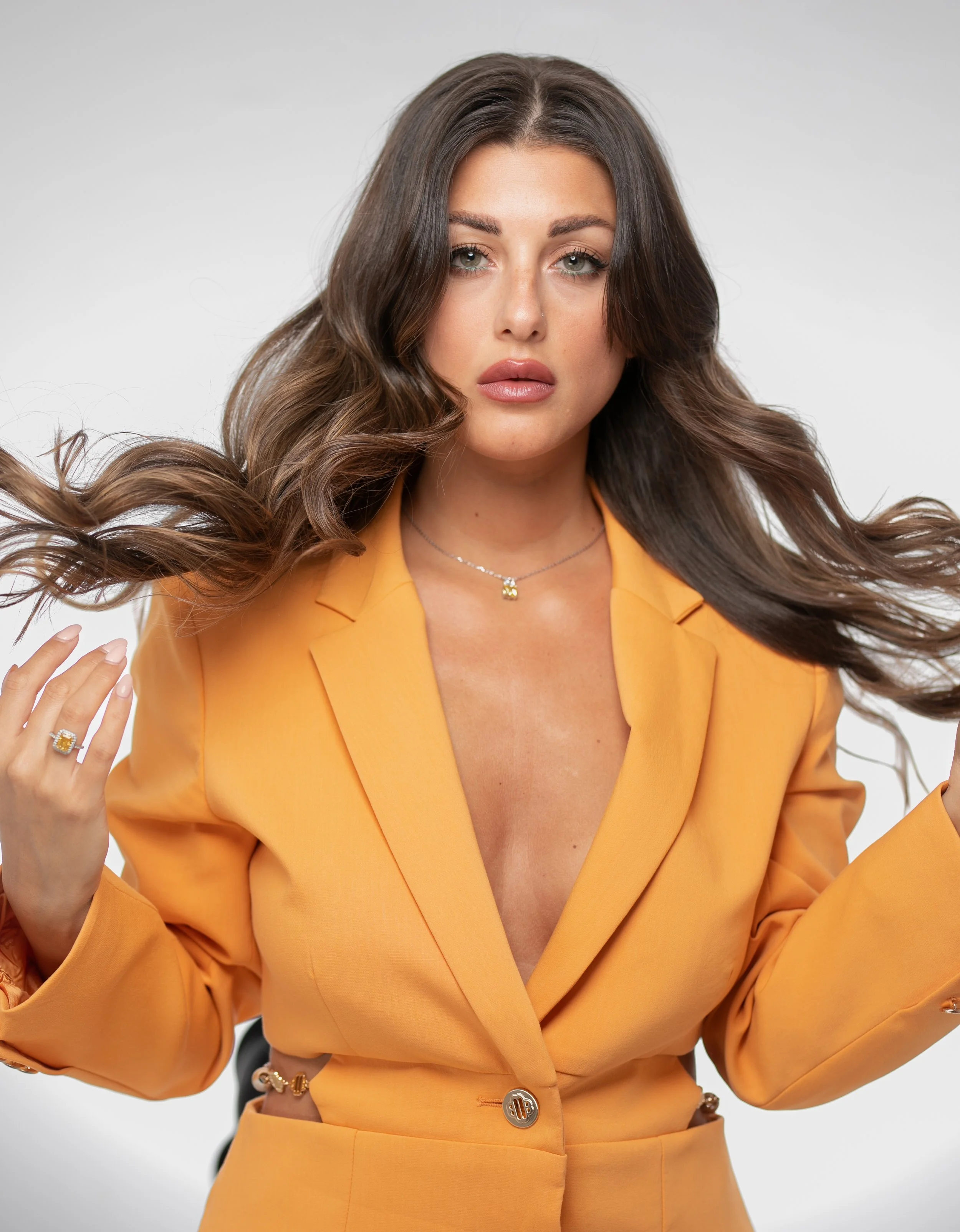 A woman with long, wavy brown hair wearing a yellow blazer and jewelry, posing in front of a gray background.