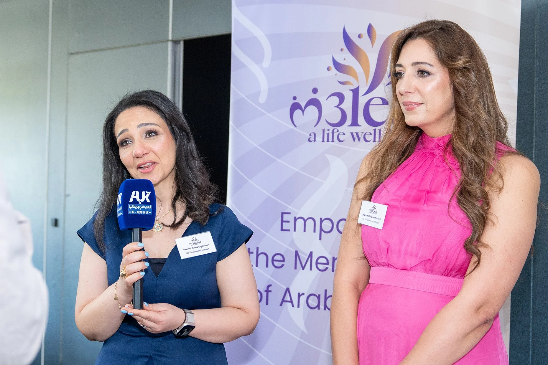 Two women at a formal event, one speaking into a microphone. The woman on the left has dark hair and is wearing a blue dress, and the woman on the right has long brown hair and is wearing a pink dress. Behind them is a banner with a logo and text, in