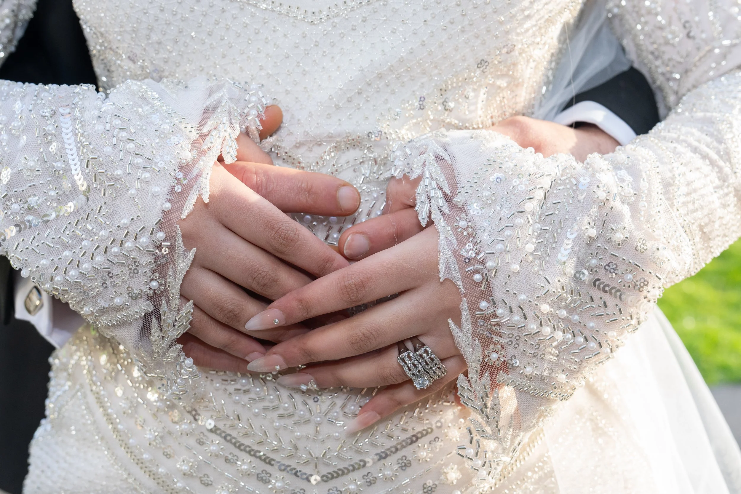 Close-up of a bride and groom's hands interlocked, showcasing the bride's wedding ring and detailed lace wedding dress with pearl and sequin embellishments.