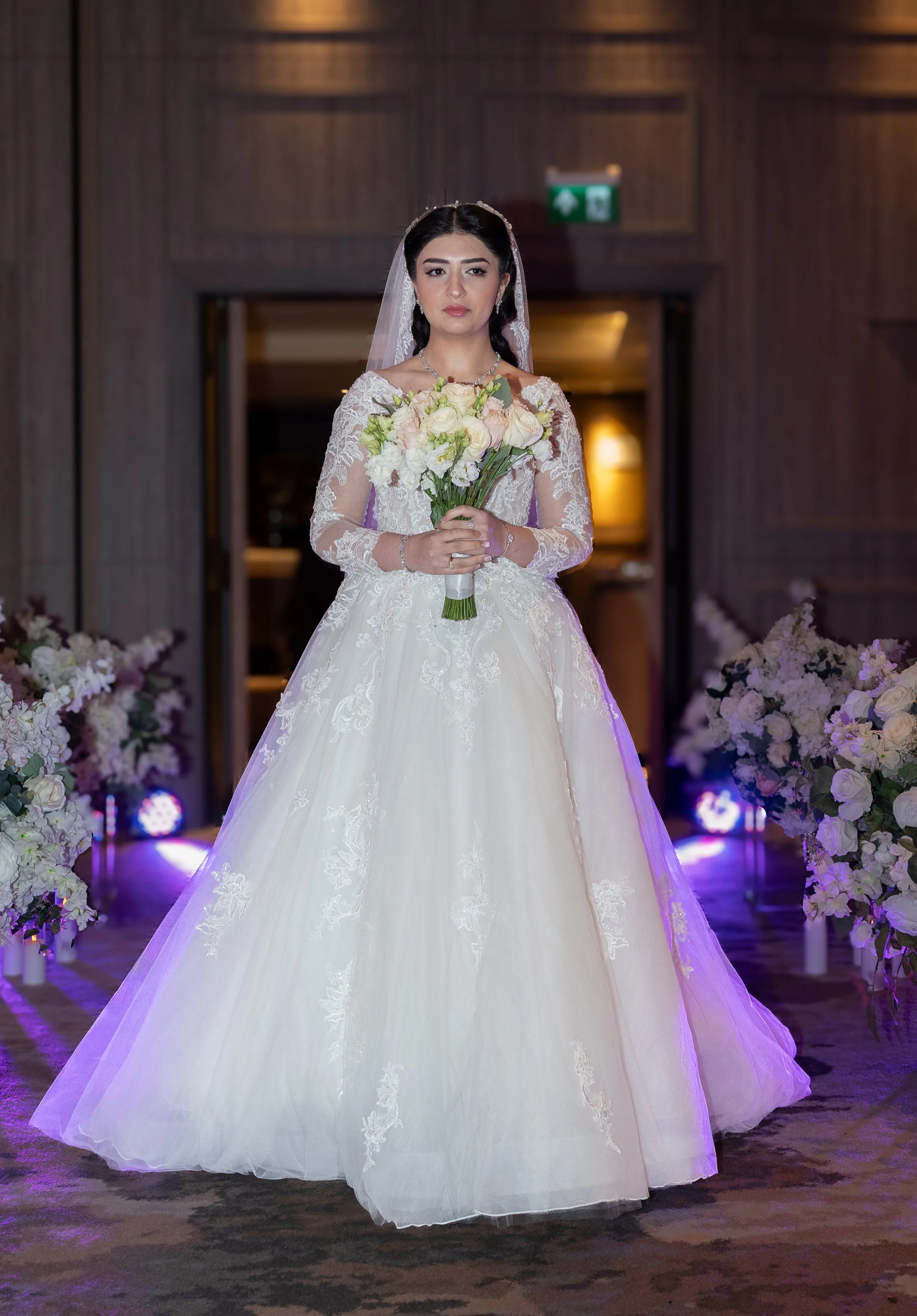 A bride standing indoors, wearing a white lace wedding gown with long sleeves, holding a bouquet of white and pink roses, and surrounded by floral decorations.