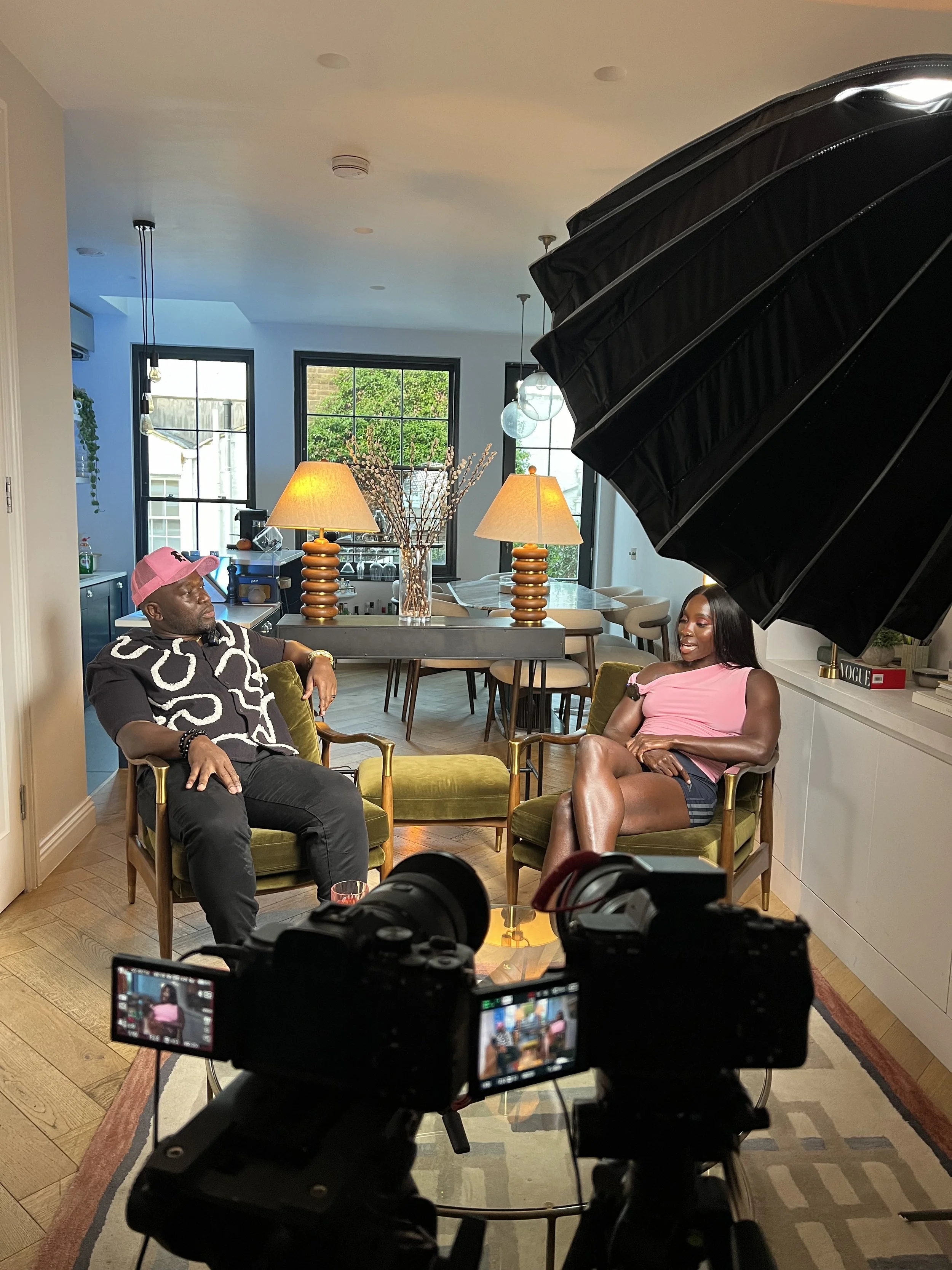 Two people having a conversation in a home setting, recorded with professional camera equipment, inside a modern dining room area with large windows, two table lamps, and decorative items.