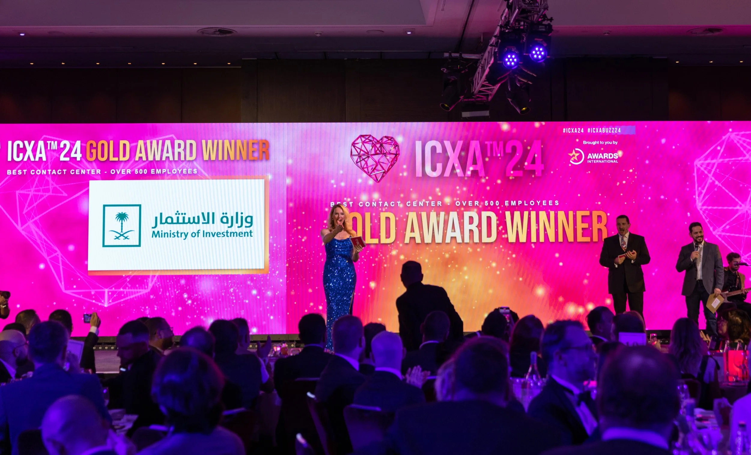 An awards ceremony with a large pink and purple digital screen displaying the text 'ICXA™ 24 Gold Award Winner' and the logo of the Ministry of Investment. Several people are on stage, including a woman in a sparkly blue dress holding a microphone an