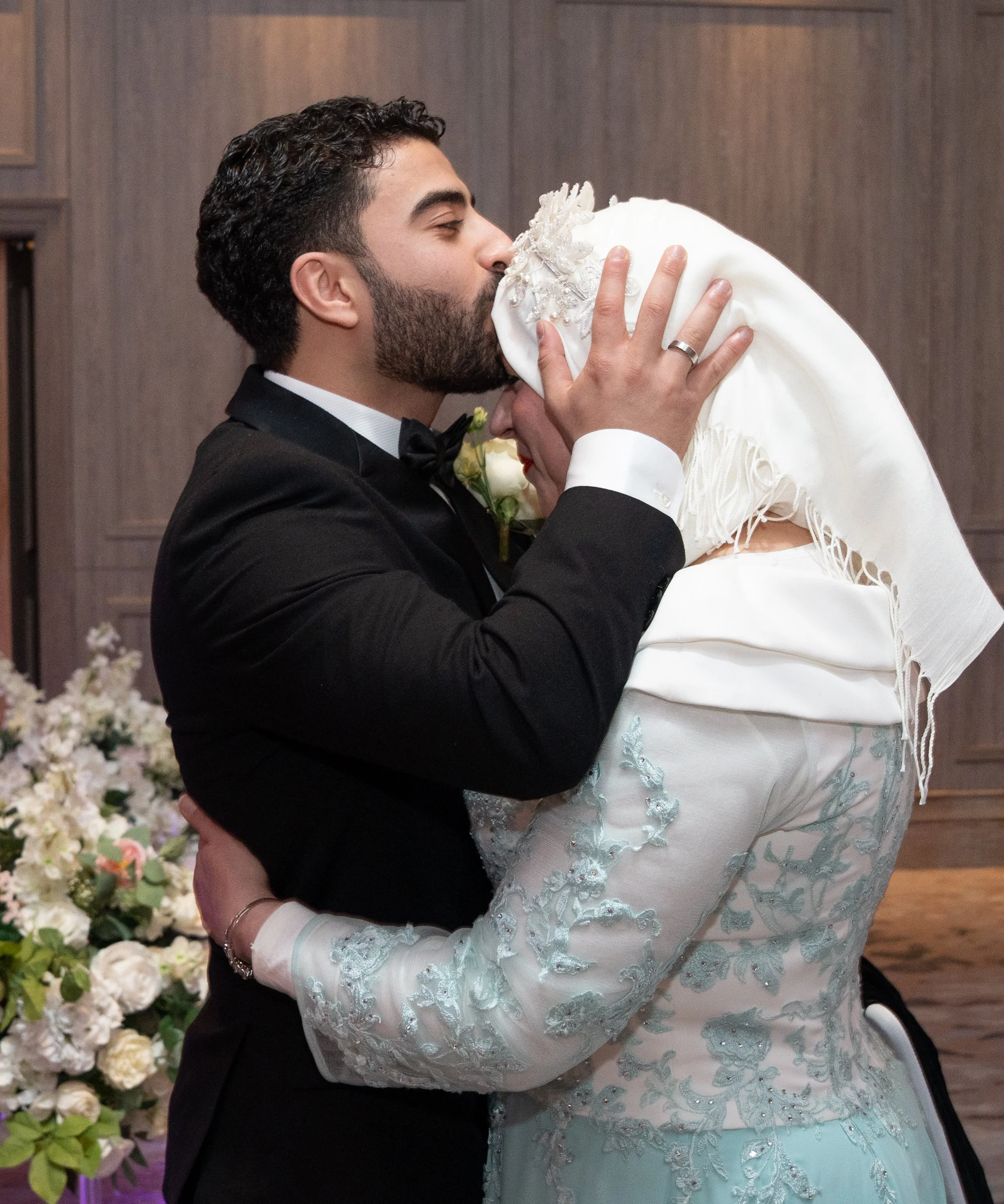 A groom in a black tuxedo kissing a bride in a white embroidered dress and headscarf during a wedding ceremony.